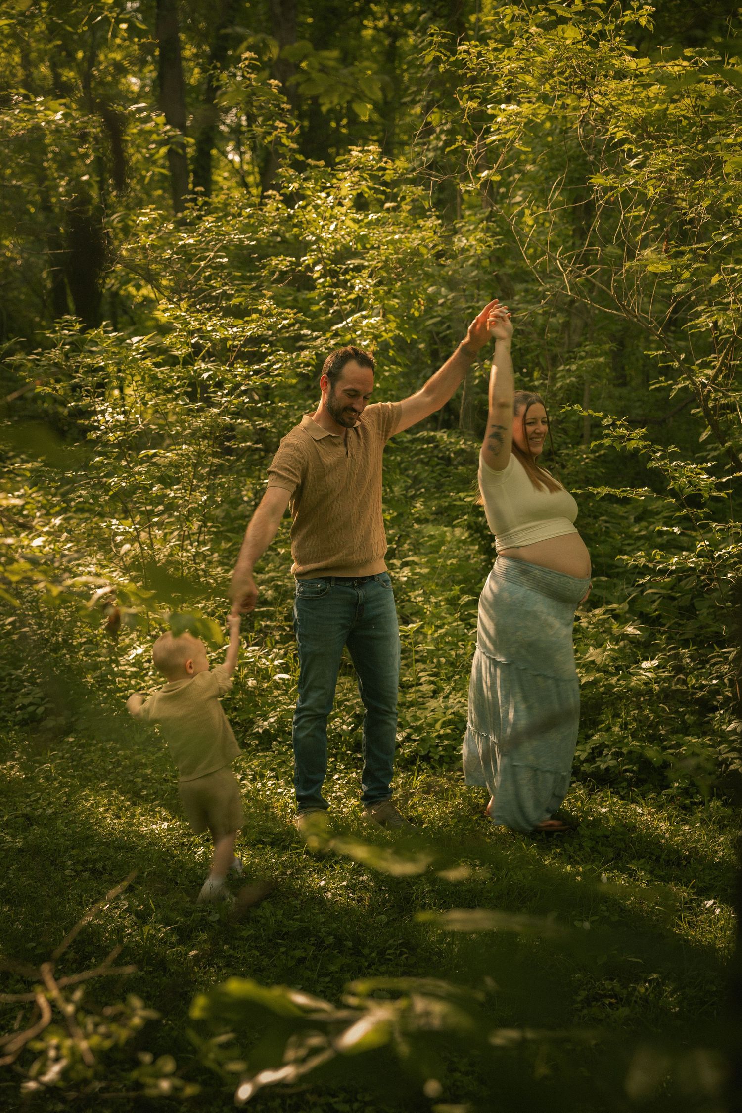 A pregnant woman dances with her husband and toddler in a lush green forest during a golden hour family photoshoot.