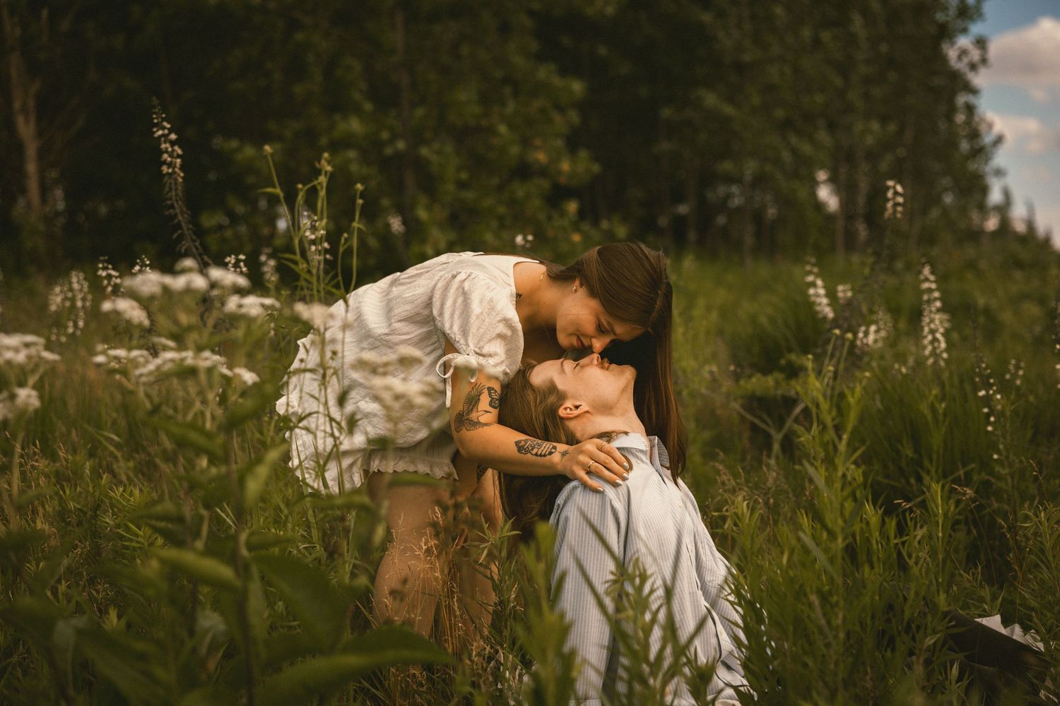 Two women sharing a tender kiss in a lush green meadow with wildflowers, captured in soft natural light.