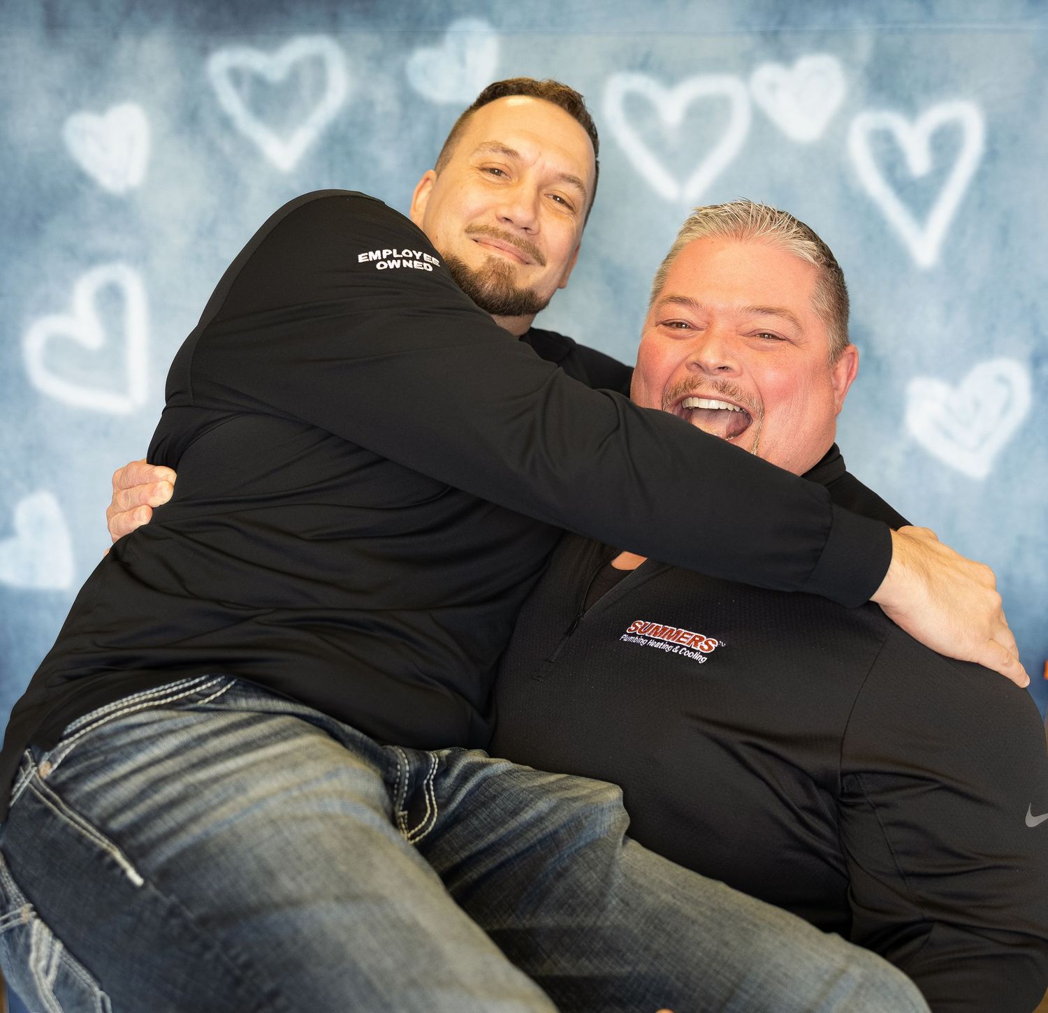 Two men in matching black company shirts share a playful hug in front of a heart-decorated backdrop.