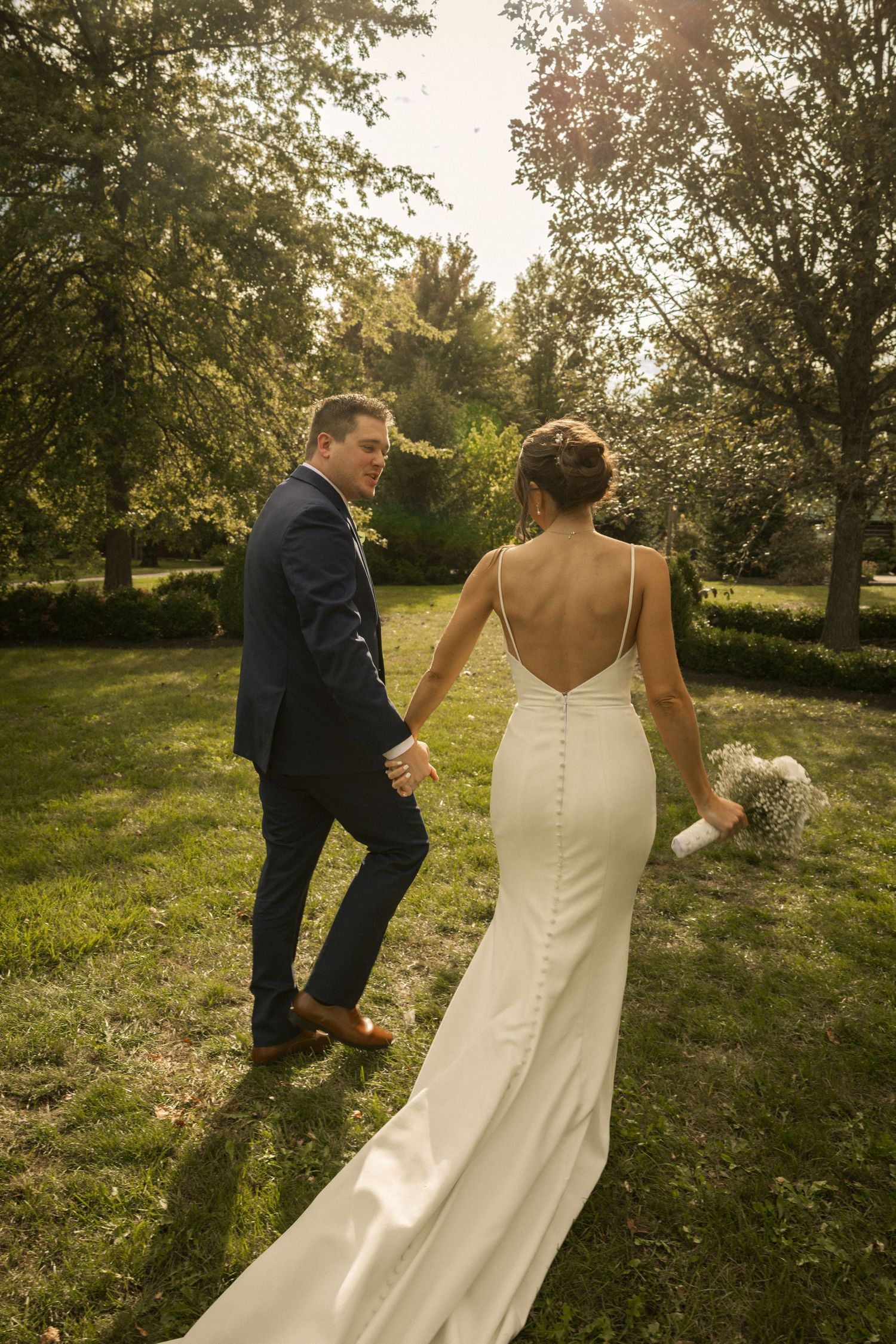 Bride and groom walk hand-in-hand through a sunlit garden, bride in backless white gown holding bouquet.