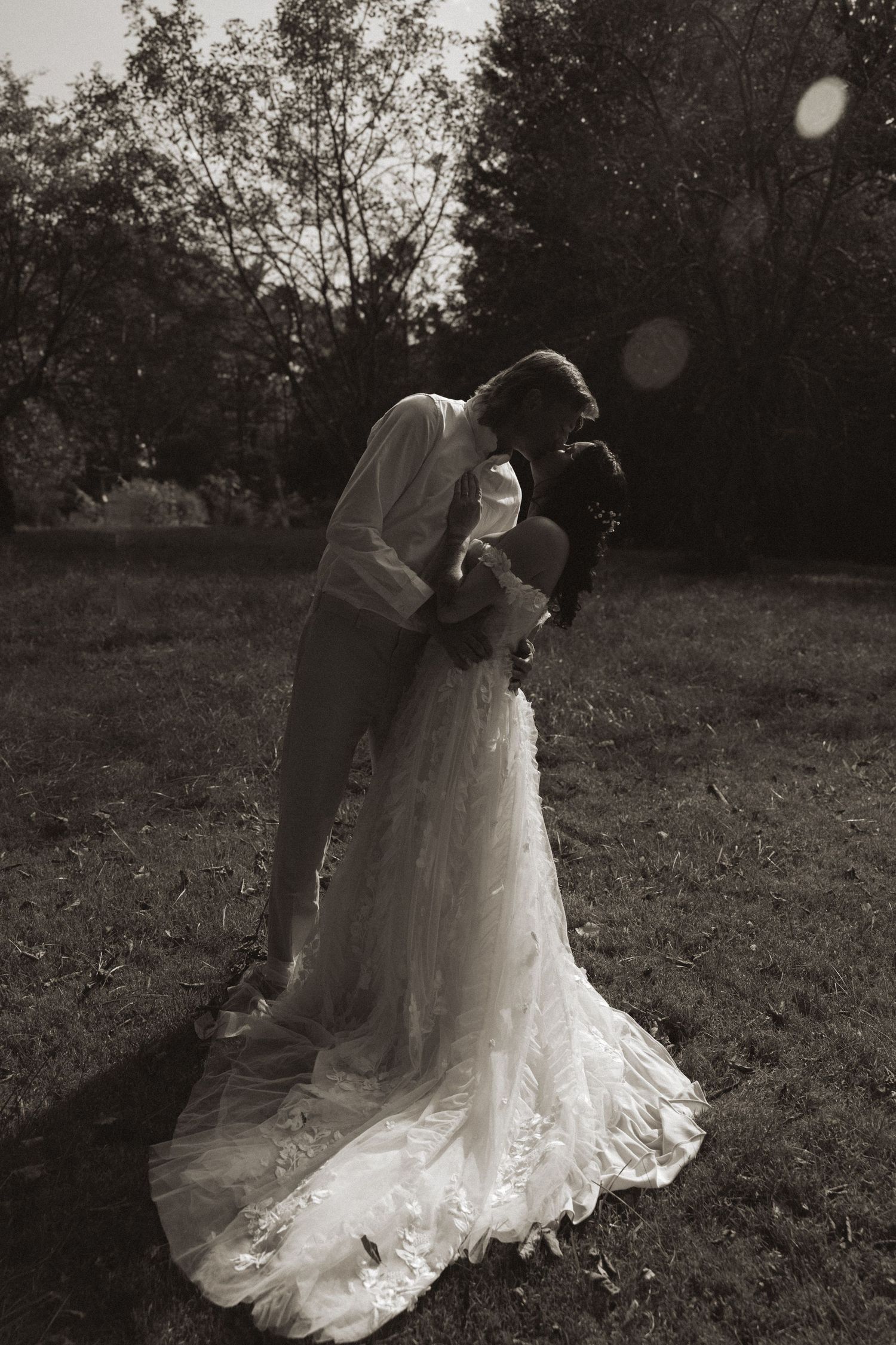 A romantic black and white photo of a couple in wedding attire embracing outdoors with a flowing lace dress train.
