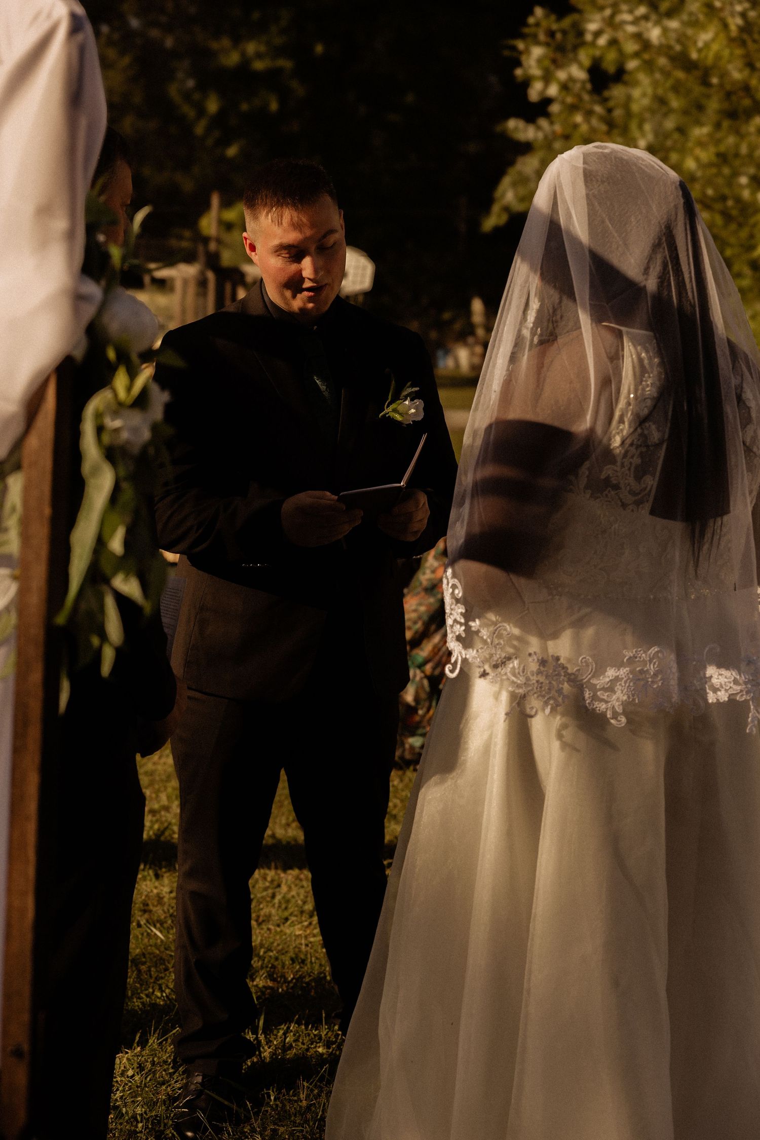 Wedding officiant reads from a book during an outdoor ceremony as bride with veil stands nearby in golden sunlight.