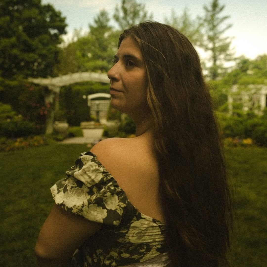 A young woman with long dark hair in a floral off-shoulder dress poses in a lush green garden.