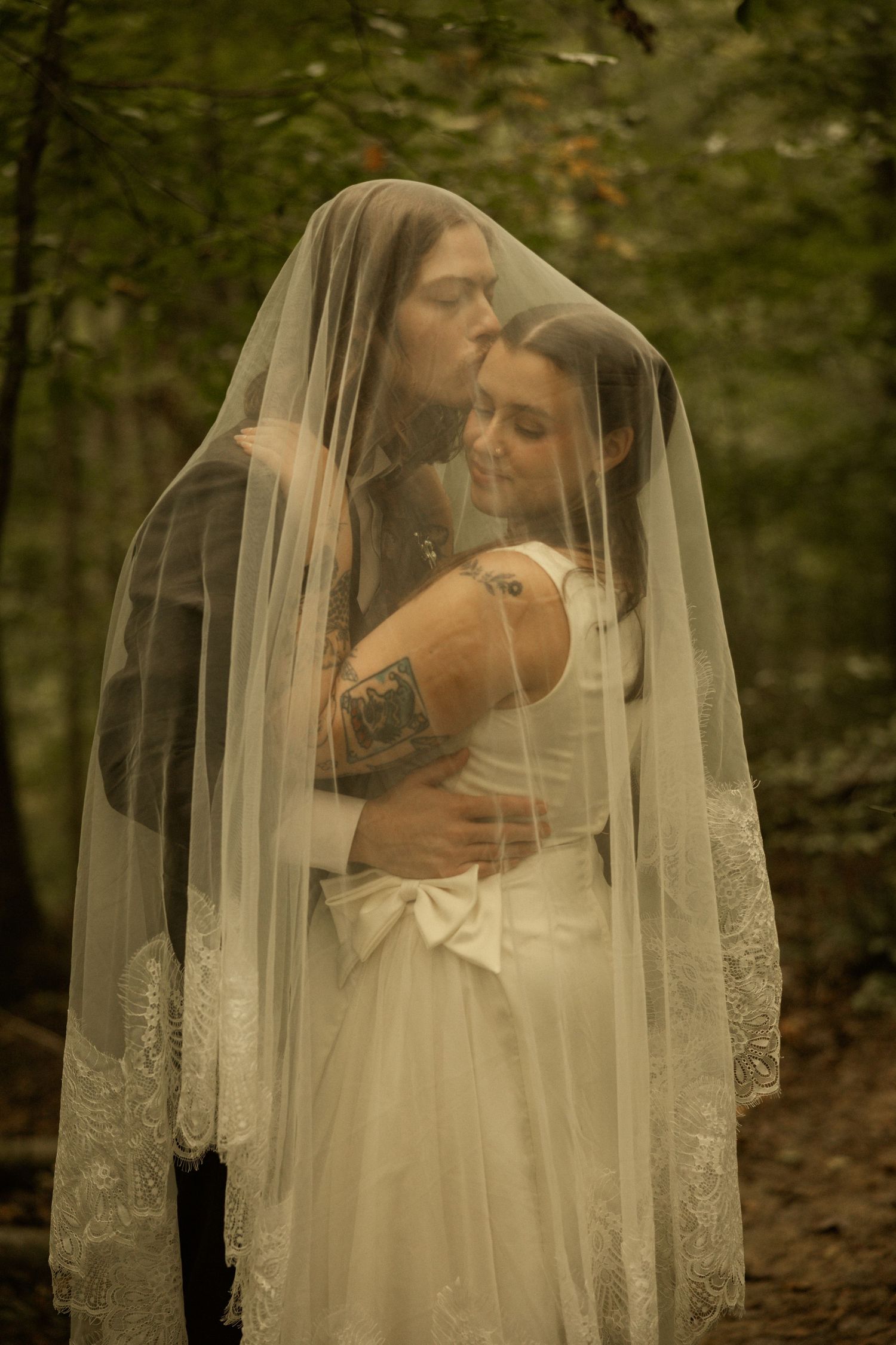 Bride and groom share a romantic embrace under a lace veil in a lush forest wedding setting.