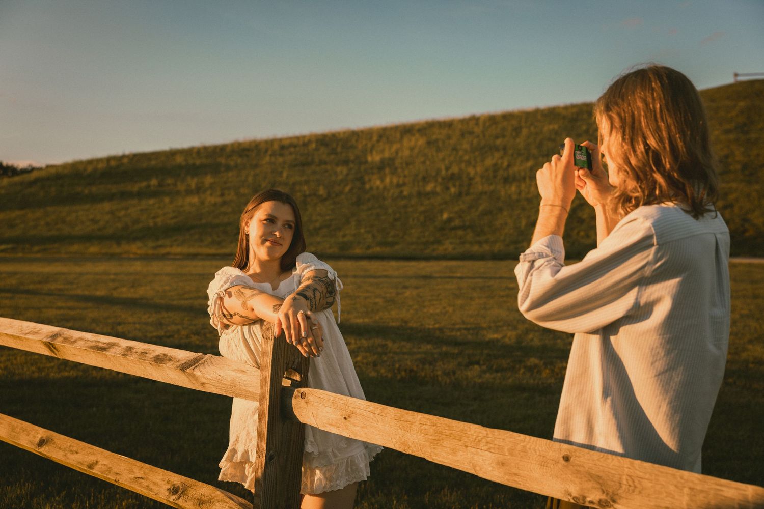 Two girls at a wooden fence in golden hour light, one posing while the other photographs her in a green field.
