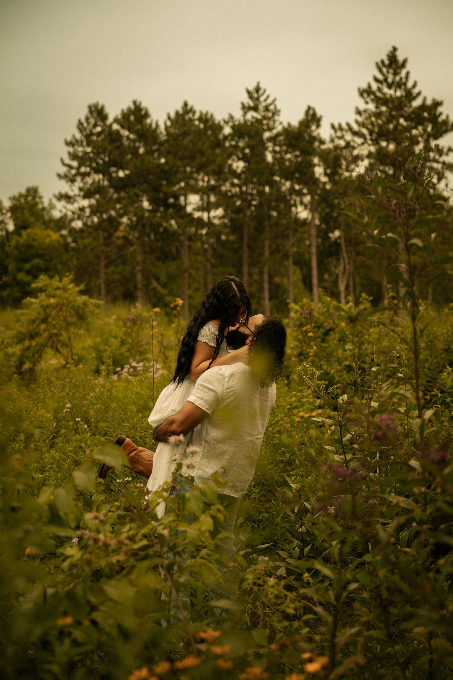 A couple embraces romantically in a lush wildflower meadow surrounded by tall pine trees on an overcast day.