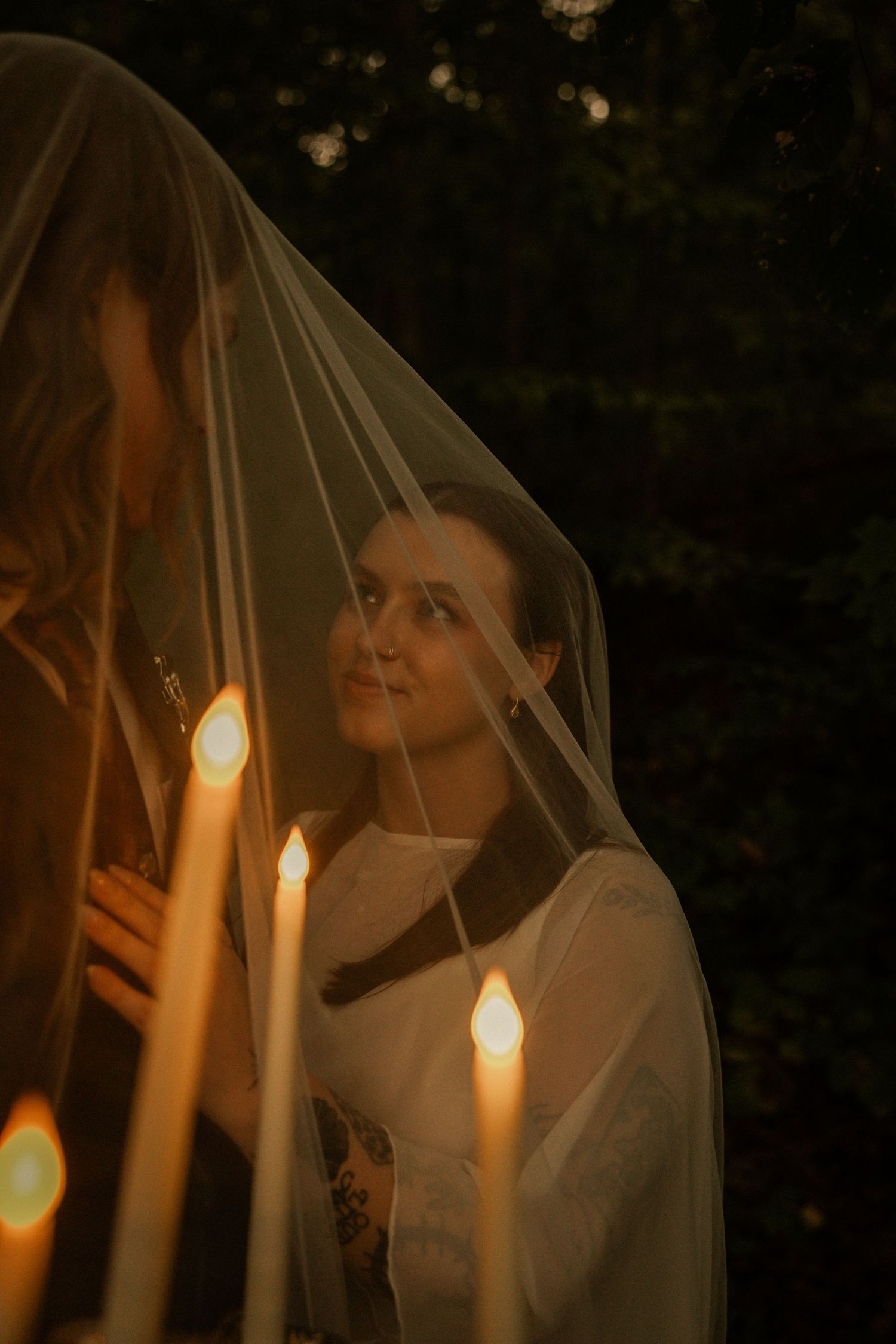 Bride smiling under a sheer veil by candlelight in a dark forest setting during a romantic wedding ceremony.