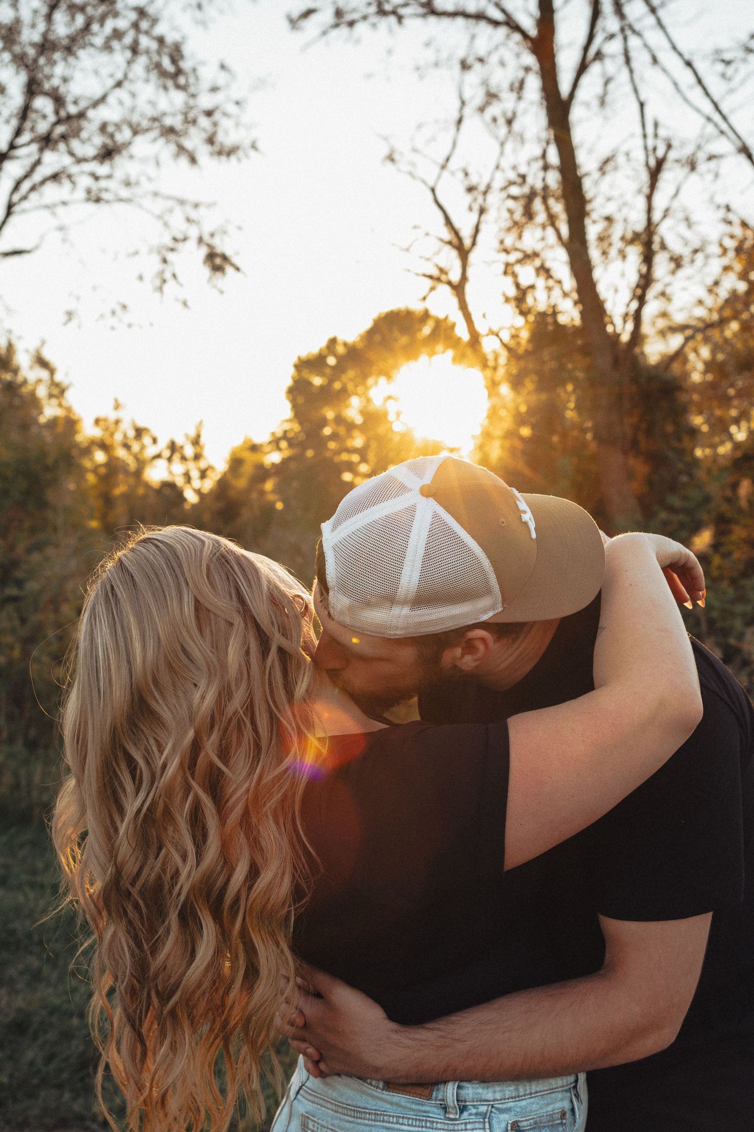 A couple embraces during golden hour with sunlit trees in the background.