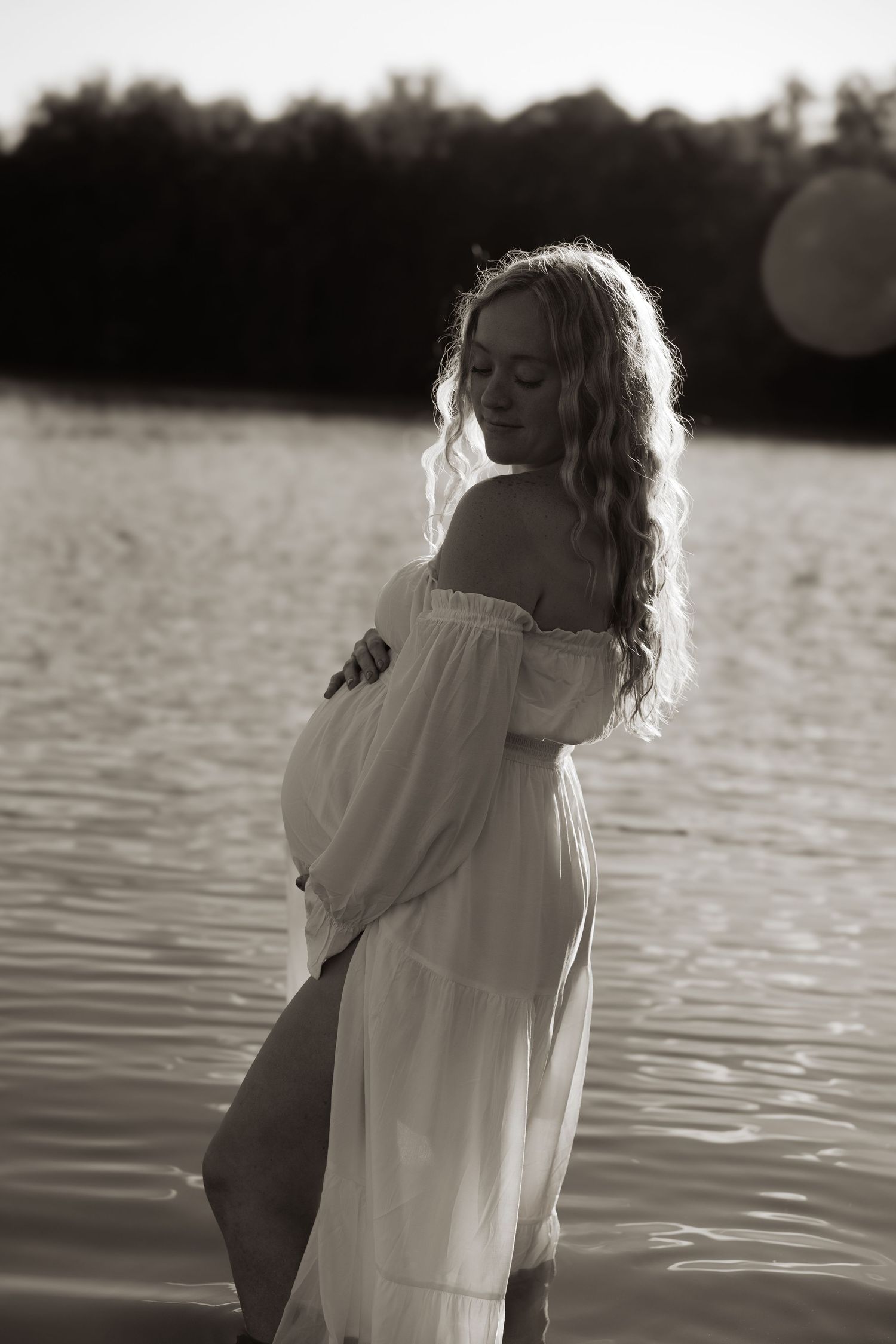 A monochrome silhouette maternity portrait series shows a person in a white shirt turning away near tranquil water at sunset.