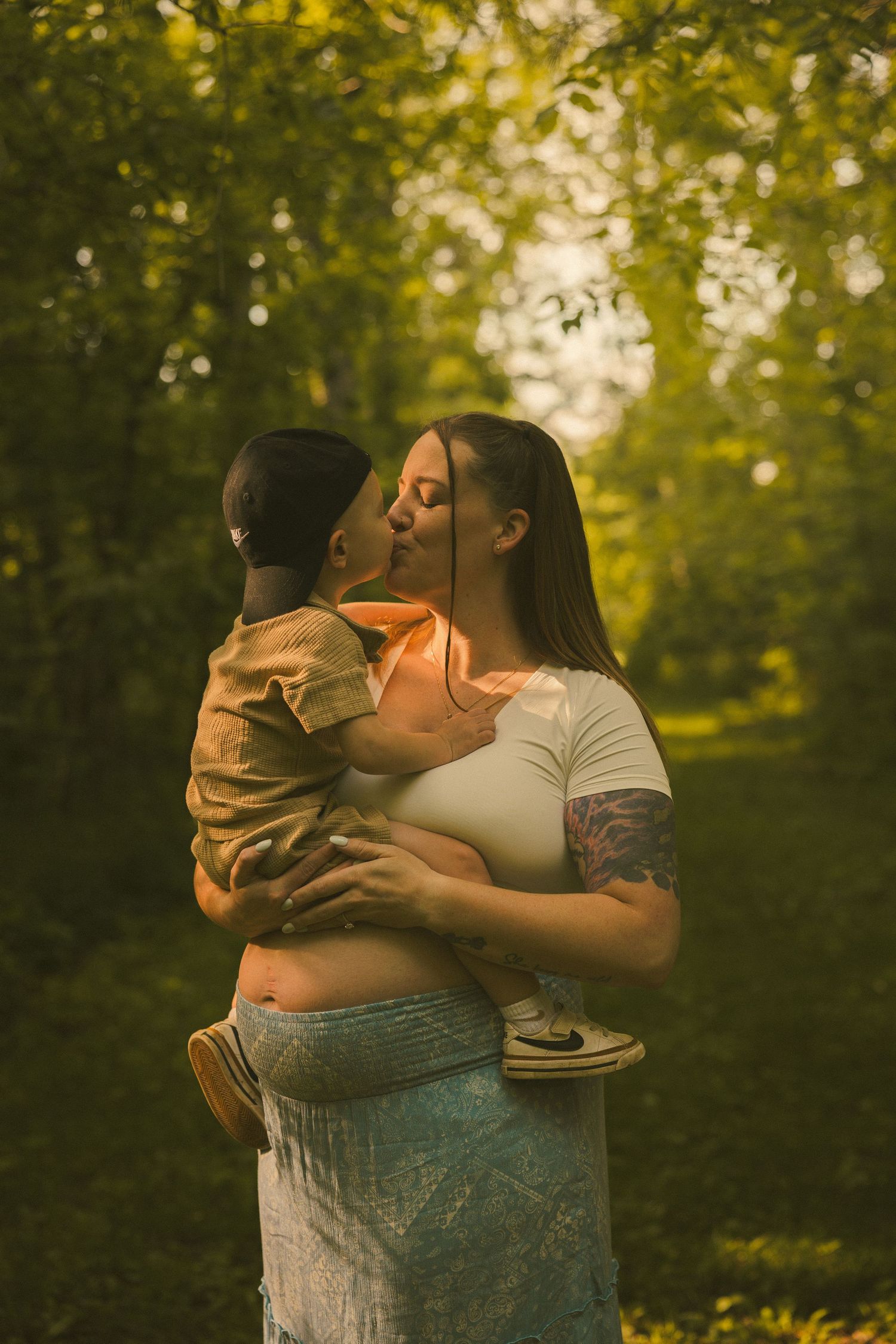 A tattooed woman kisses a young child she holds in her arms outdoors in golden sunlight among green trees.