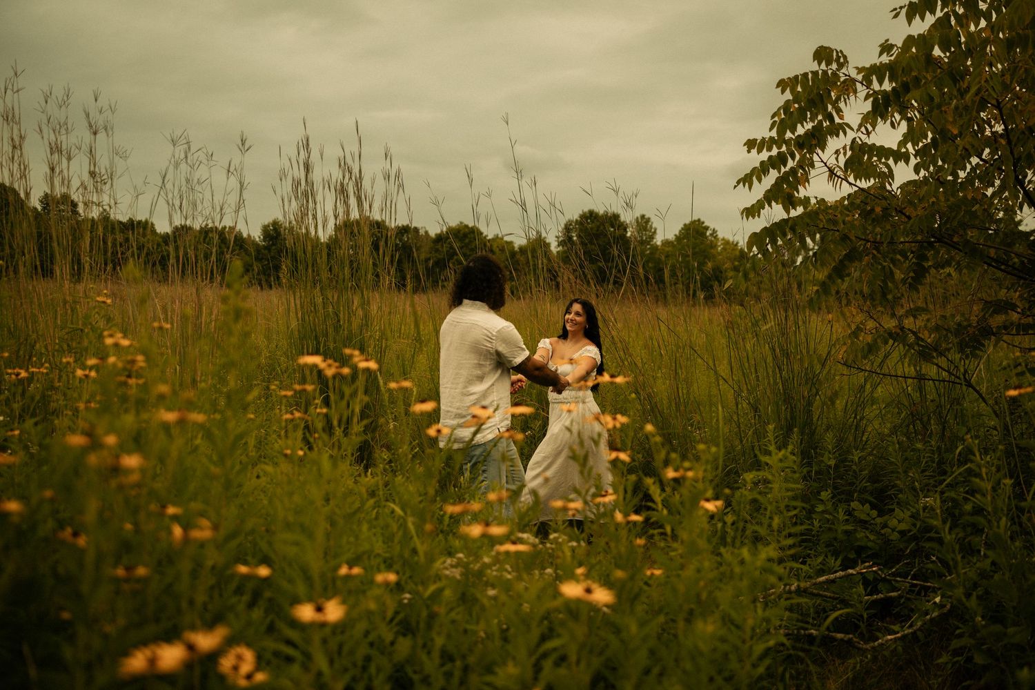 Couple in white outfits standing in a wildflower meadow with tall grass and orange flowers under cloudy skies.