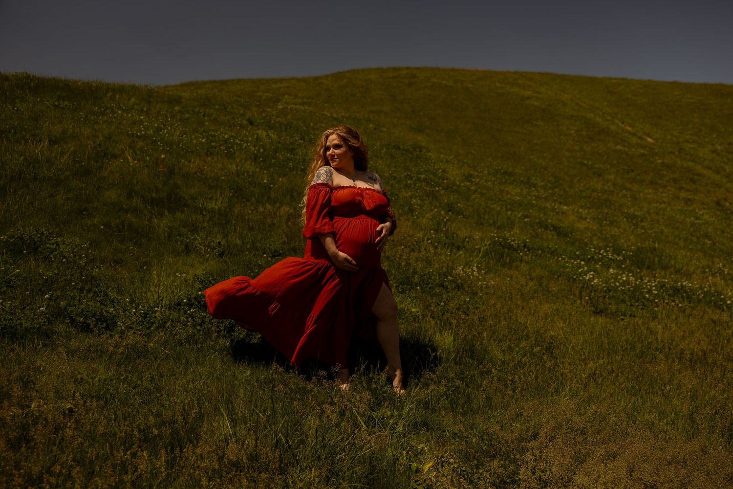 A dramatic portrait of someone in a flowing red dress standing on a grassy hillside.