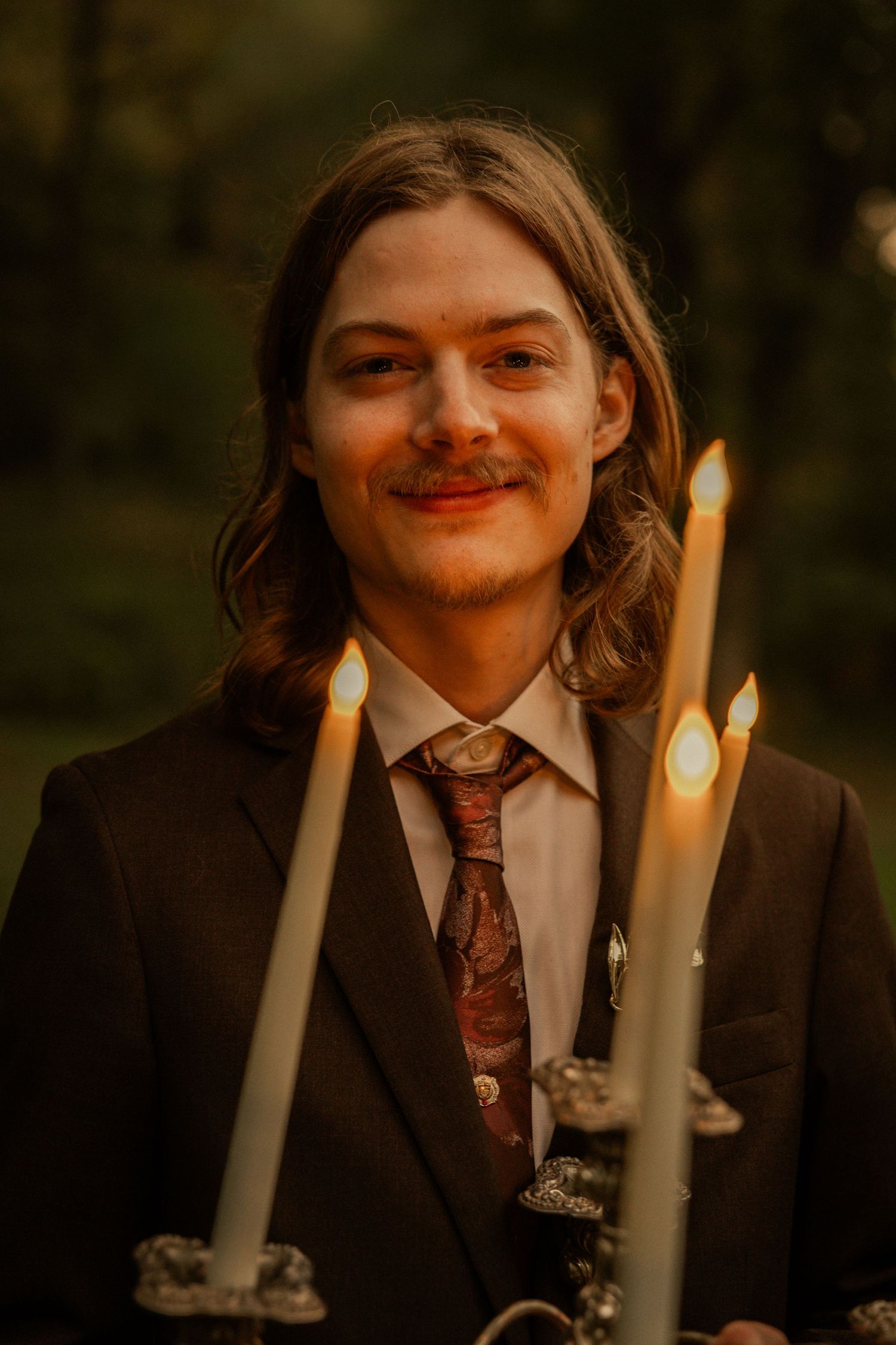 Young man with long hair and mustache in dark suit holding lit candelabra outdoors at dusk.
