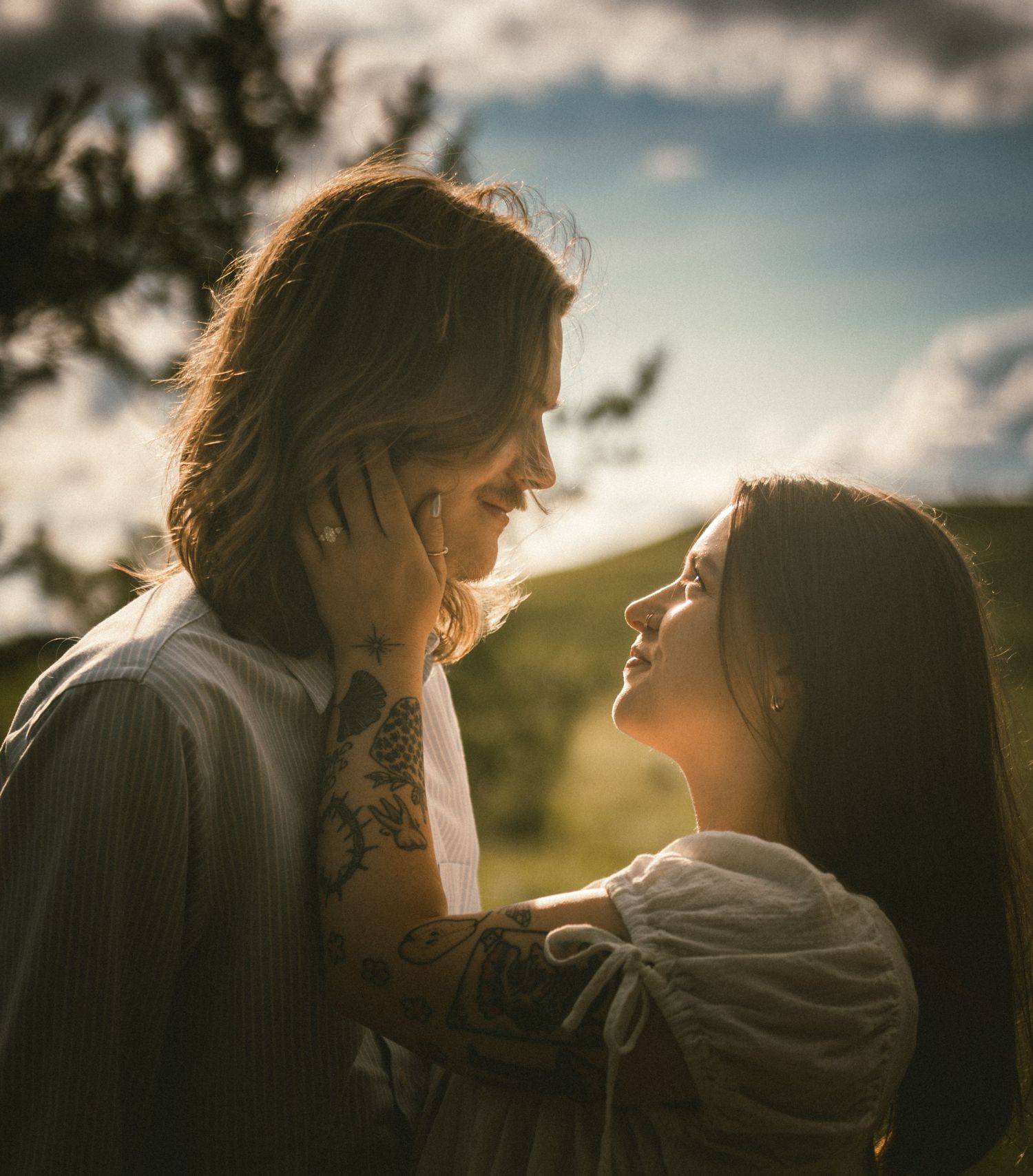 A tattooed couple shares a romantic moment outdoors at golden hour, gazing into each other's eyes with soft sunlight behind them.