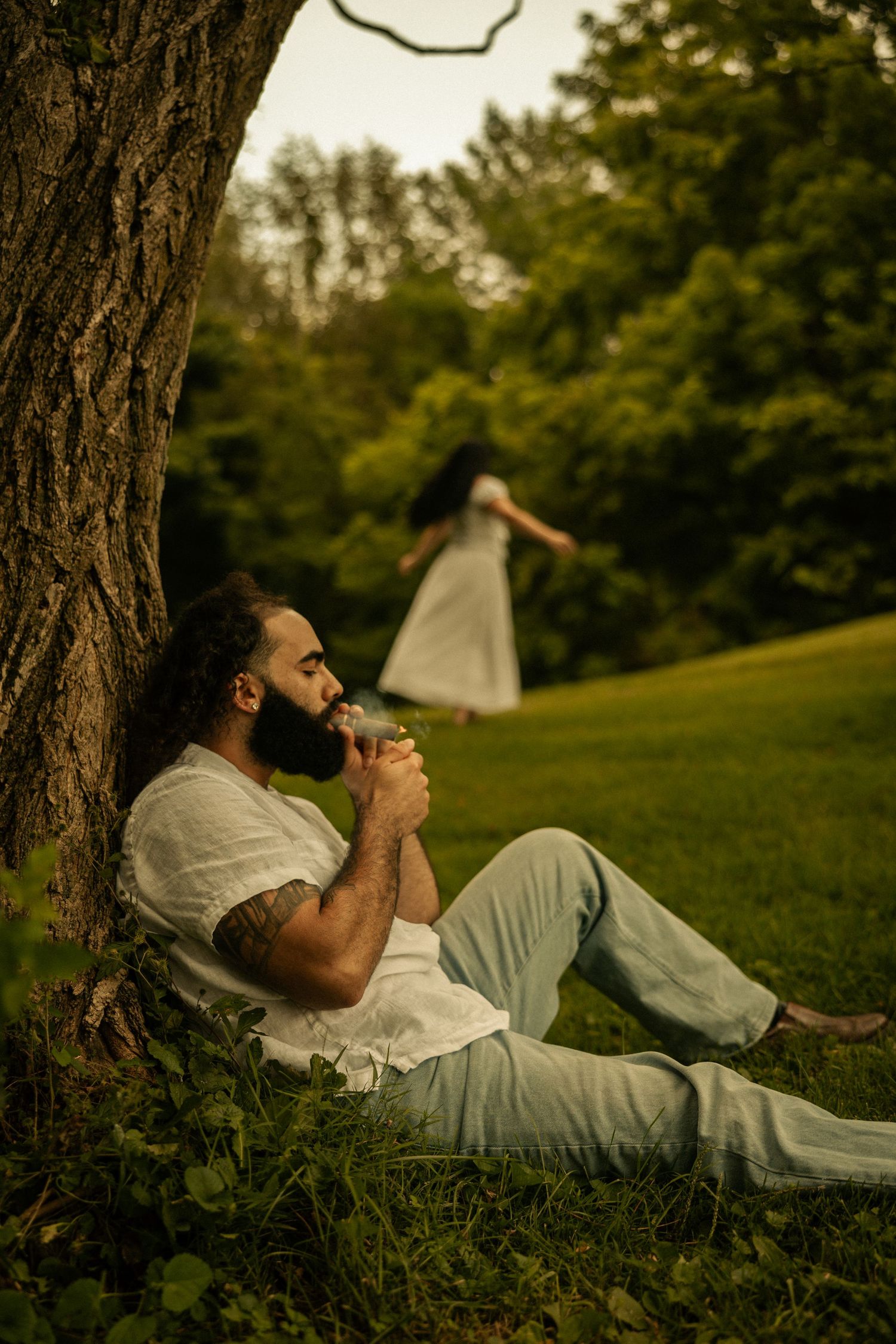 A bearded man with tattoos reclines against a tree in a park, while a woman in a white dress stands in the background.