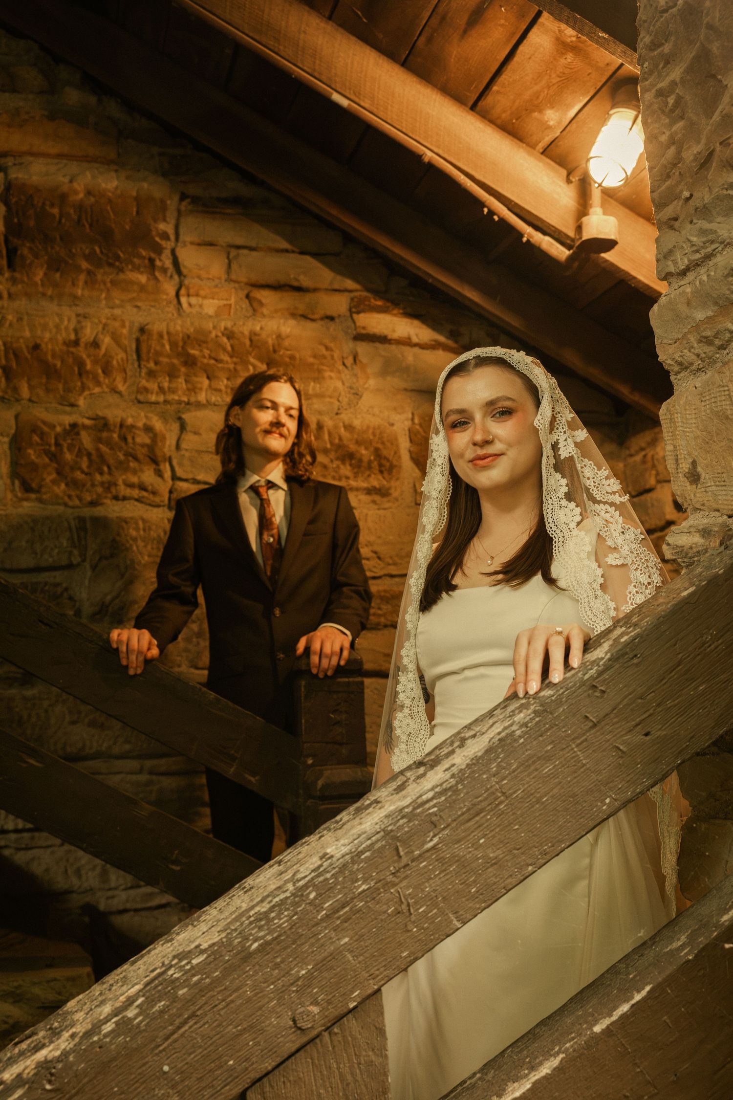Bride in lace veil and groom in dark suit pose on rustic stone staircase in warm candlelit setting.