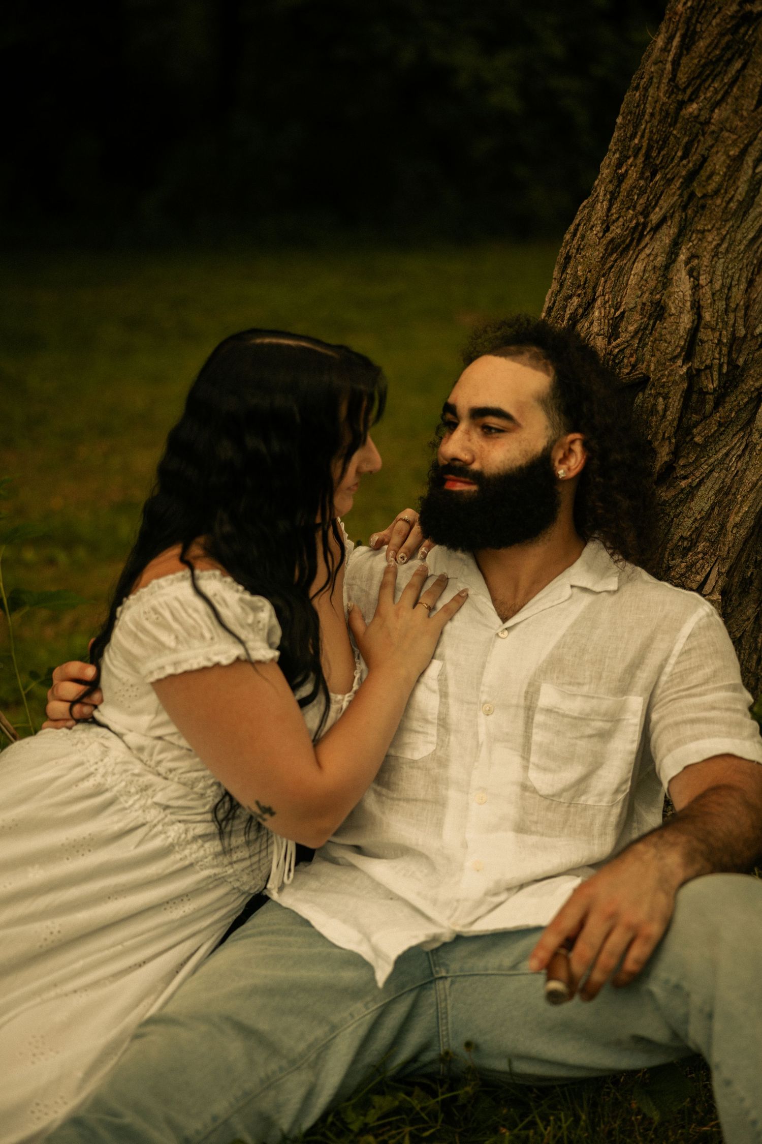 A bearded man in white linen shirt sits against a tree as a woman in white dress tenderly touches his face outdoors.