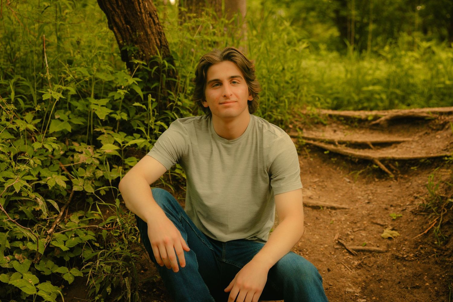 Young man in gray t-shirt and jeans sitting outdoors in a lush green forest setting.