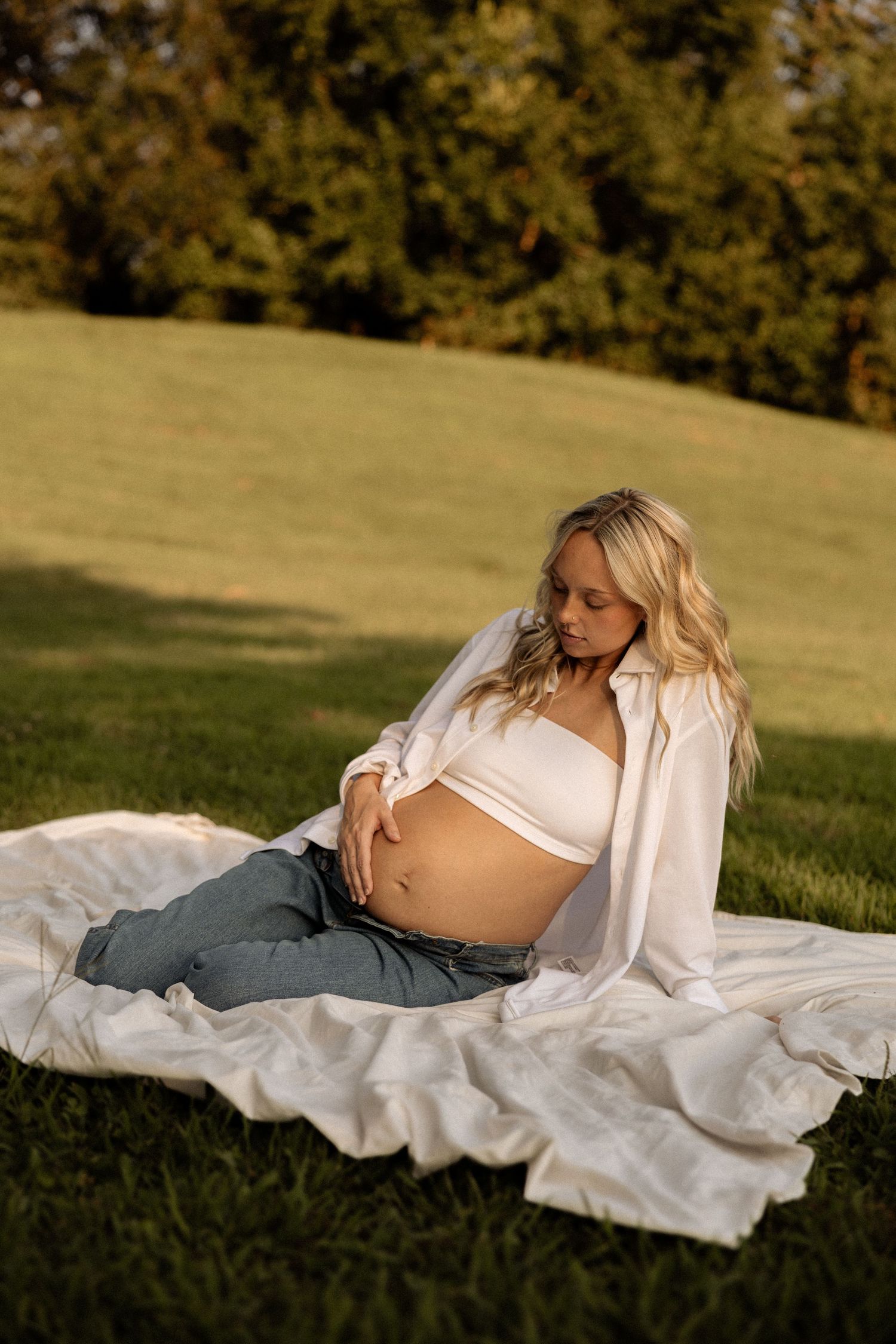 Expectant mother reclines on white fabric during outdoor maternity session at sunset.