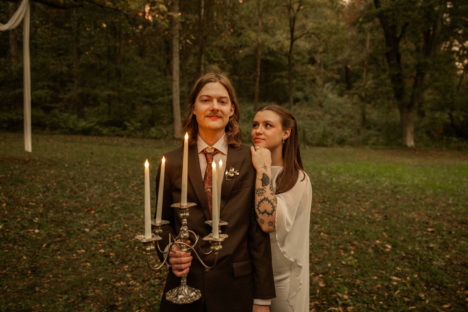 Two women pose outdoors at a woodland wedding, one holding an elegant silver candelabra with lit candles.
