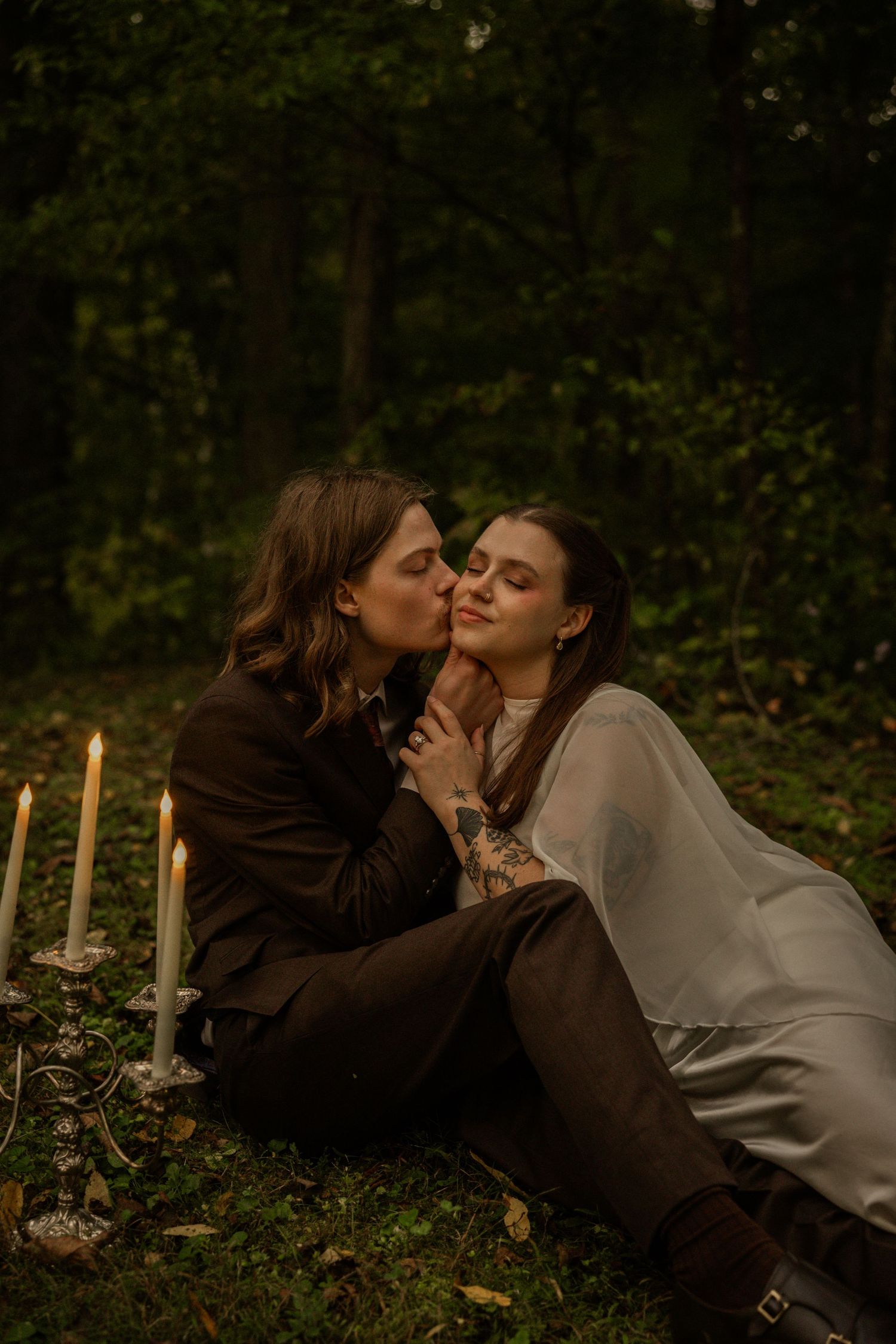 Couple sharing a romantic kiss in a dark forest setting, surrounded by candlelight on grass.