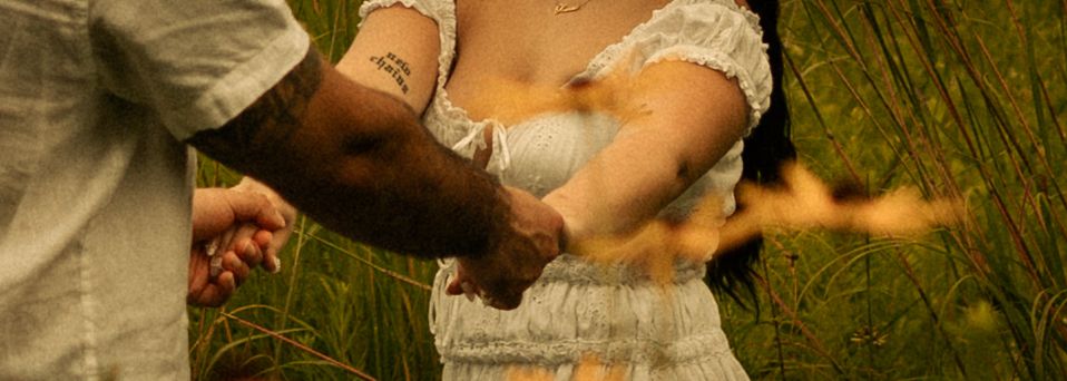 A couple holds hands in a sunlit field, the man's tattooed arm visible as they walk together through golden wildflowers.