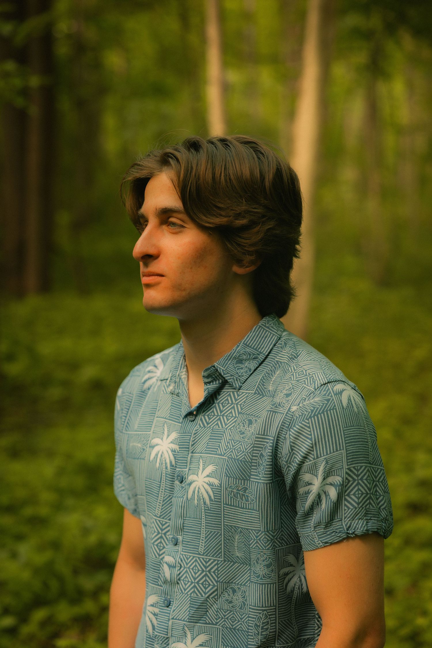 Young man in blue Hawaiian shirt standing in lush green forest, looking thoughtfully to the side.