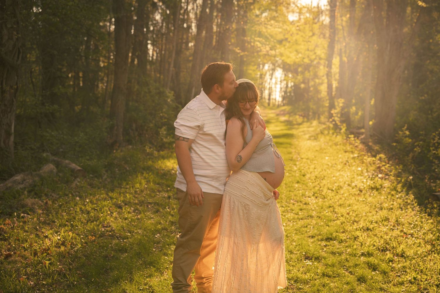 Couple sharing a tender moment in a sunlit forest path, man kissing pregnant woman in a flowing white dress.