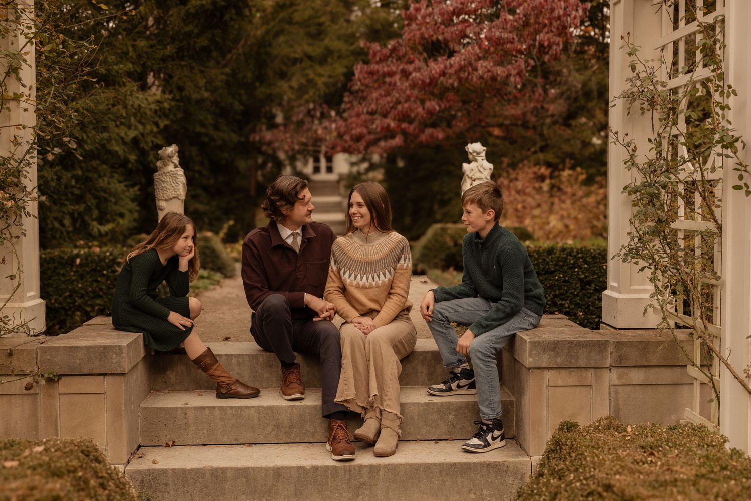 A group sits together in a romantic garden pergola with autumn foliage and stone columns during golden hour.