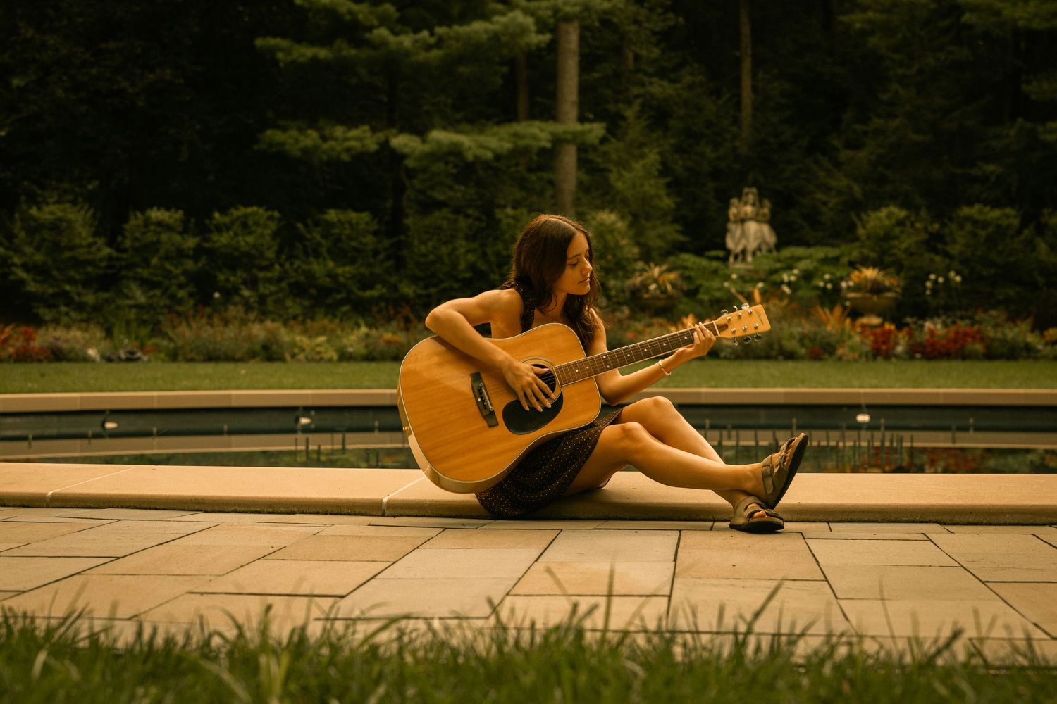 Young woman playing acoustic guitar while sitting on stone pavers beside a pool, surrounded by lush green trees.