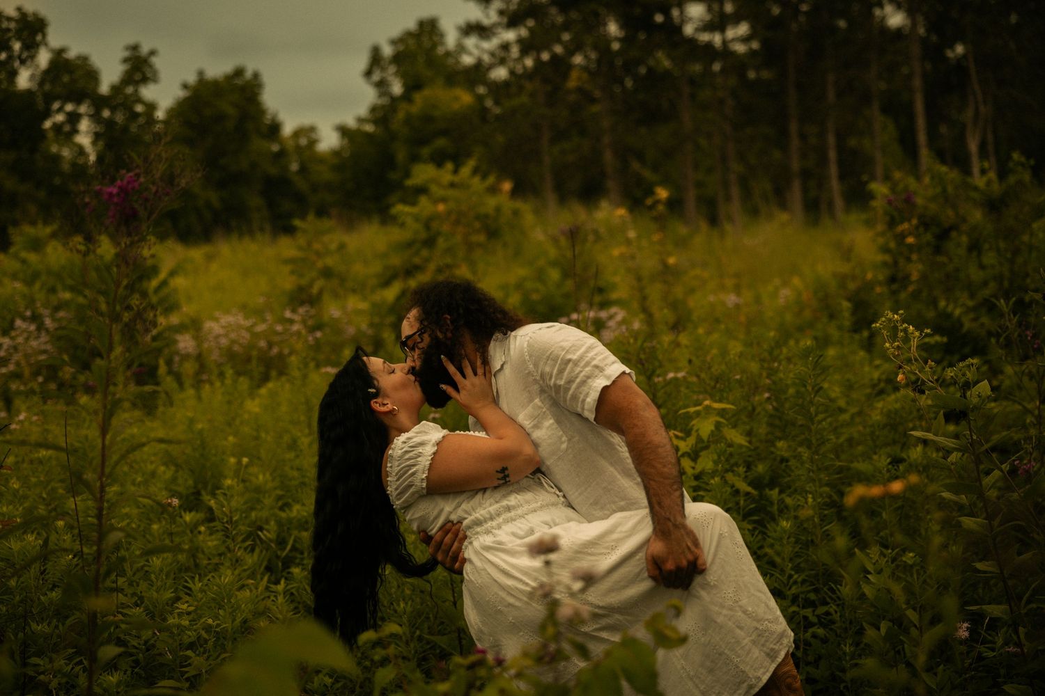 Couple sharing a romantic kiss in a lush green meadow surrounded by wildflowers and trees.