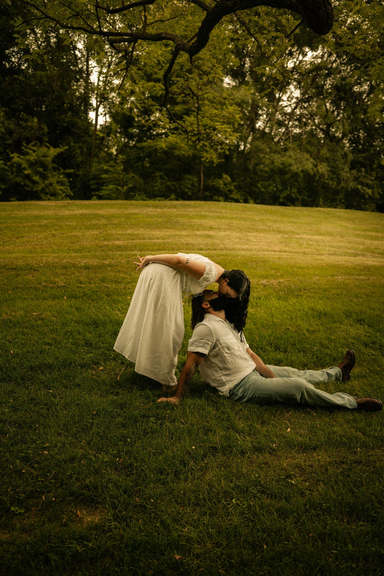 A couple shares a romantic kiss on a lush green lawn, woman in white dress bending to kiss man seated on grass.