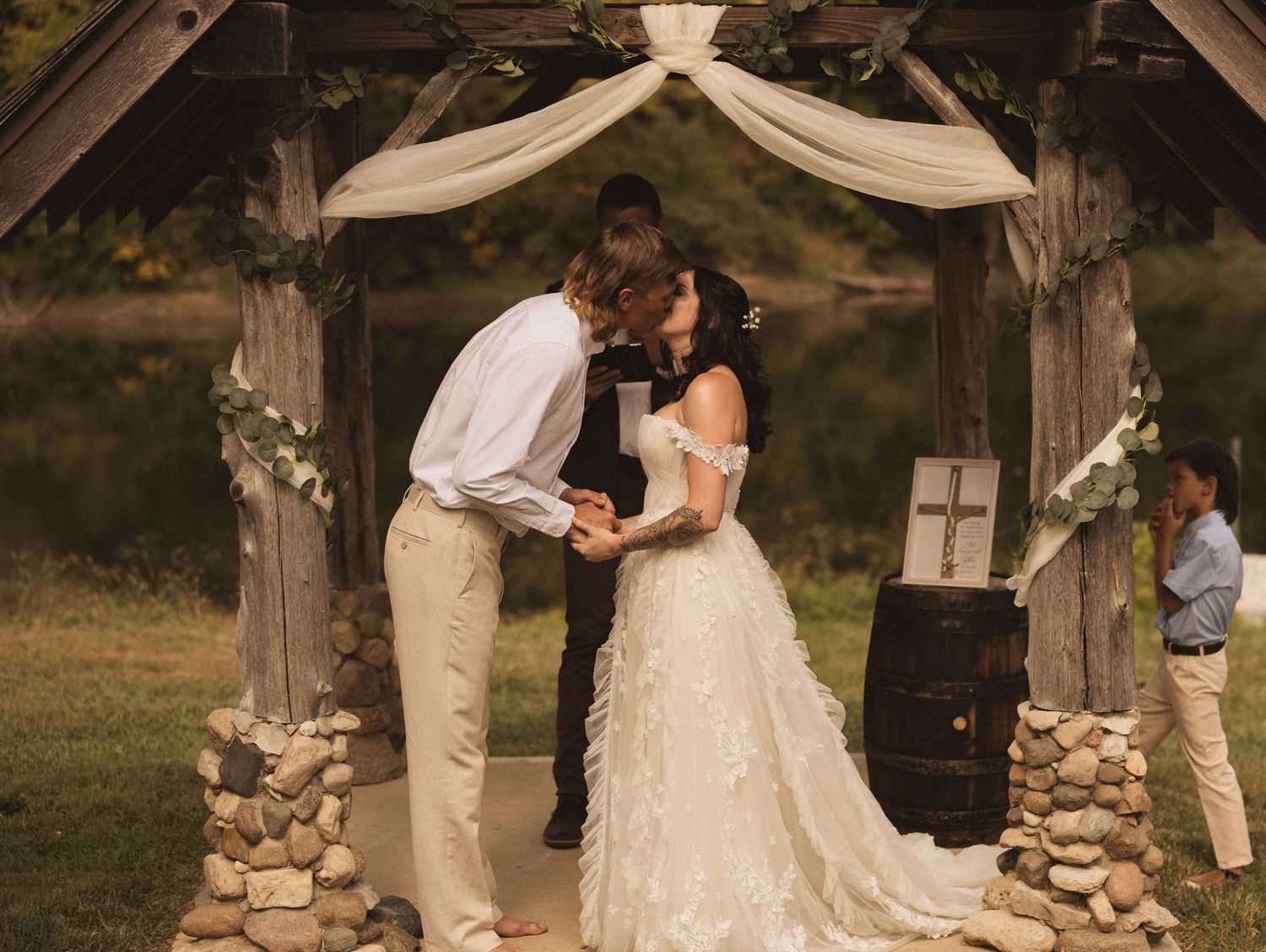 A romantic wedding ceremony takes place under a rustic wooden arch decorated with white fabric and floral arrangements.