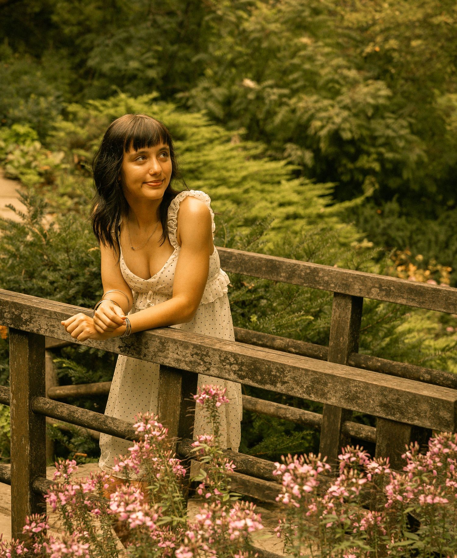 Young woman in white sundress leans on wooden bridge railing surrounded by lush greenery and pink flowers.