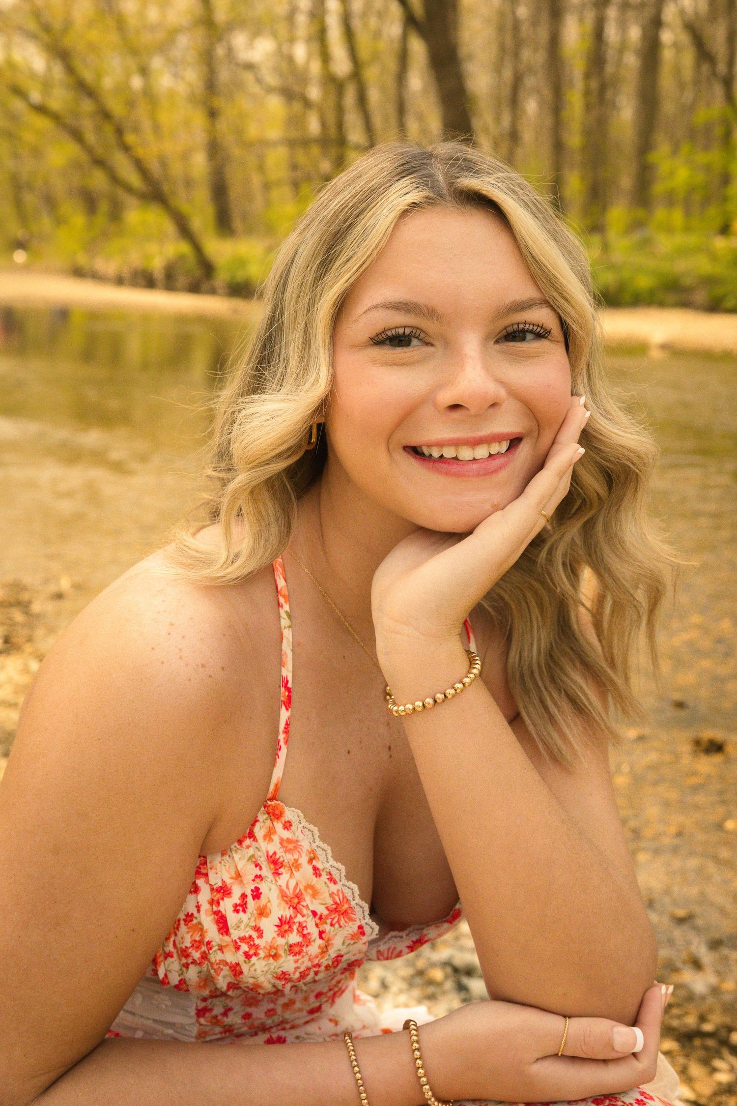 Smiling blonde woman in floral dress poses outdoors by a forest pond, resting chin on hand.