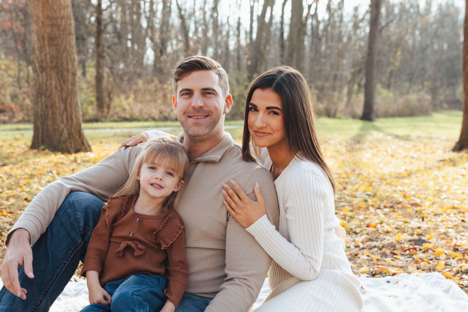 Family of three posing together on fallen autumn leaves in neutral tones.