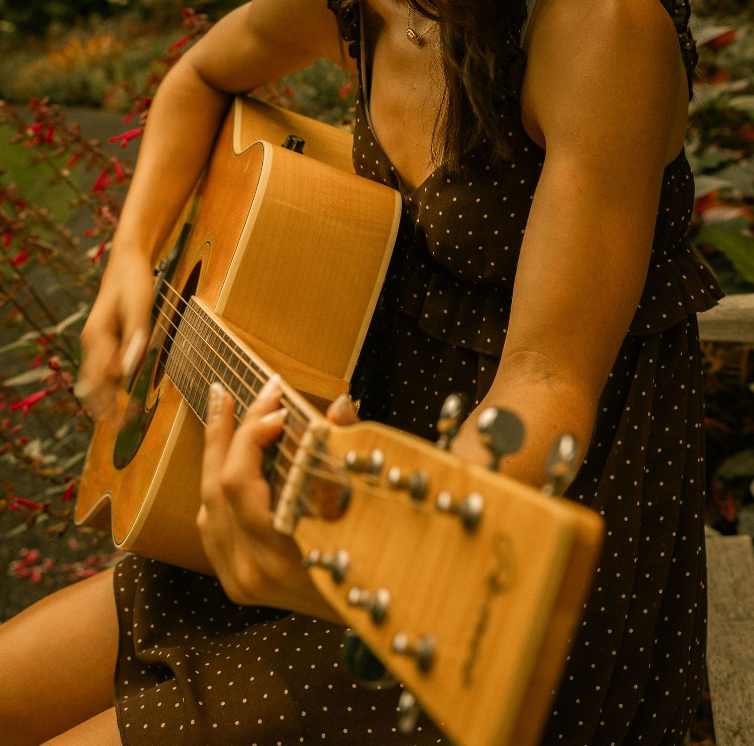 Woman in polka dot dress playing acoustic guitar outdoors in a garden setting.