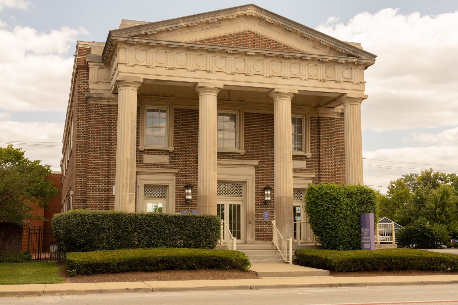 Neoclassical brick building with white columns and portico, featuring manicured hedges and steps leading to the entrance.