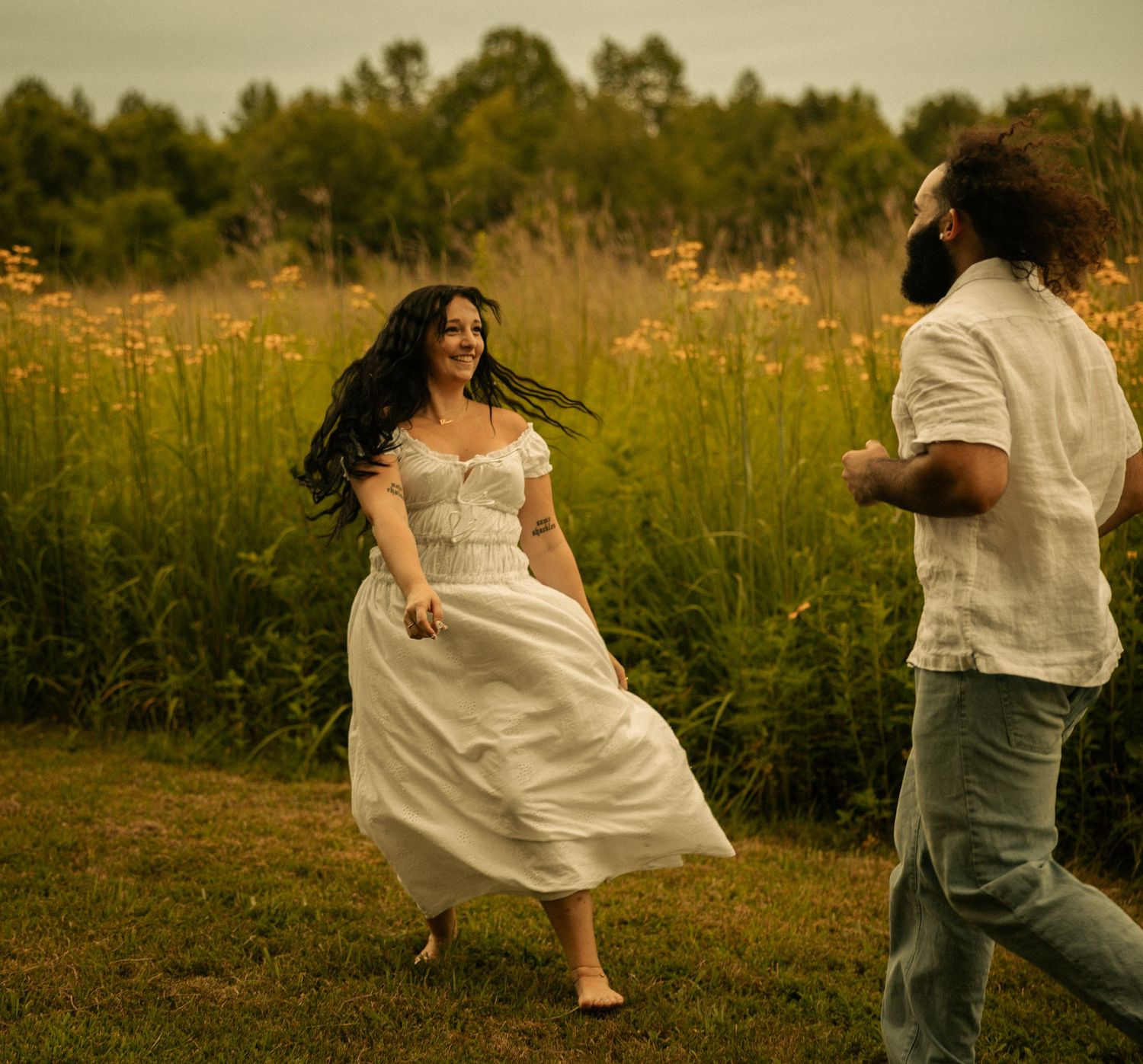 A joyful couple runs through a lush meadow, the woman in a flowing white dress, surrounded by wildflowers.