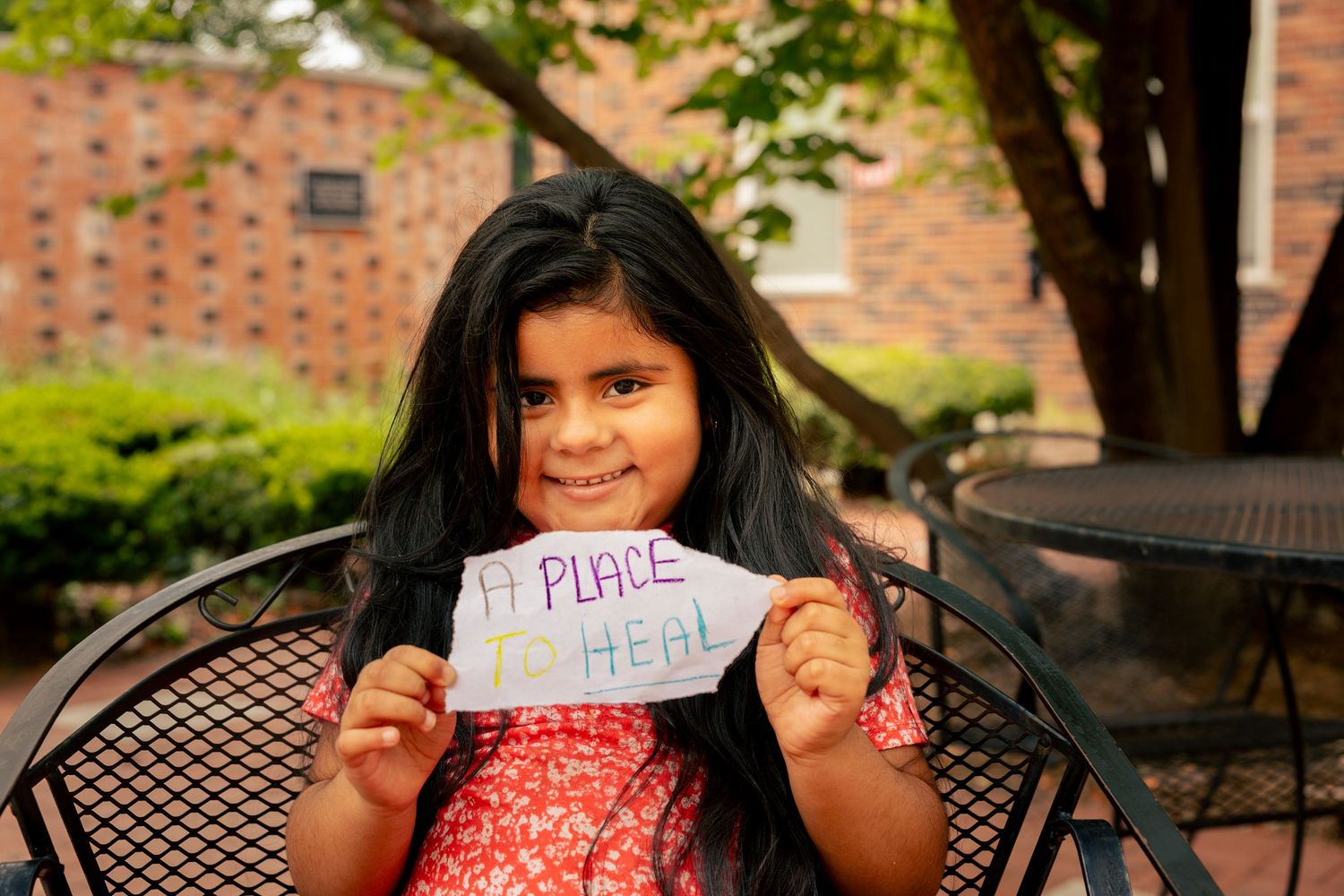 Young girl smiling outdoors holding a handwritten sign reading A Place to Heal, seated at a patio table.