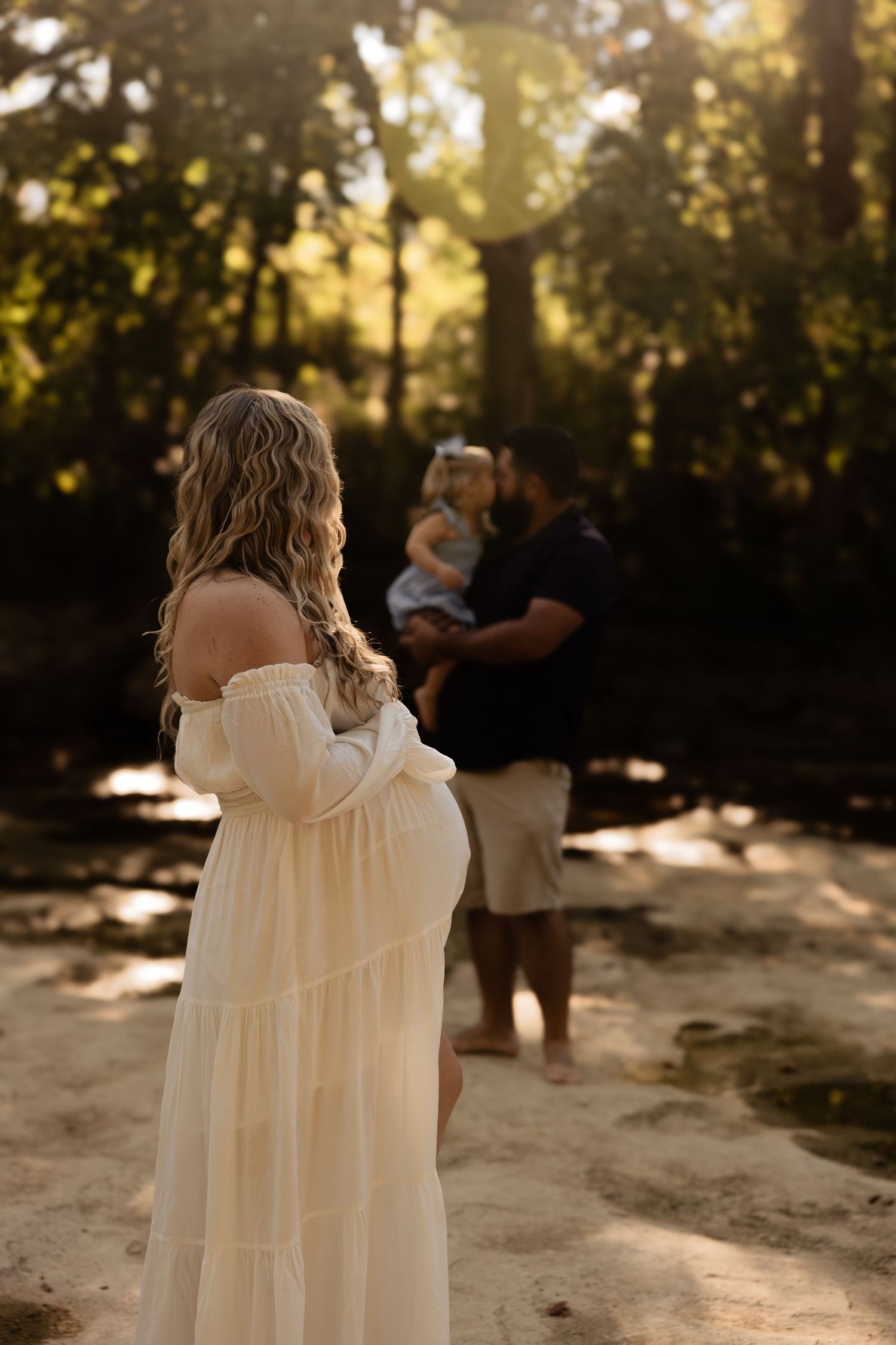 Figure in a flowing cream-colored dress stands in dappled sunlight against a wooded backdrop during golden hour.