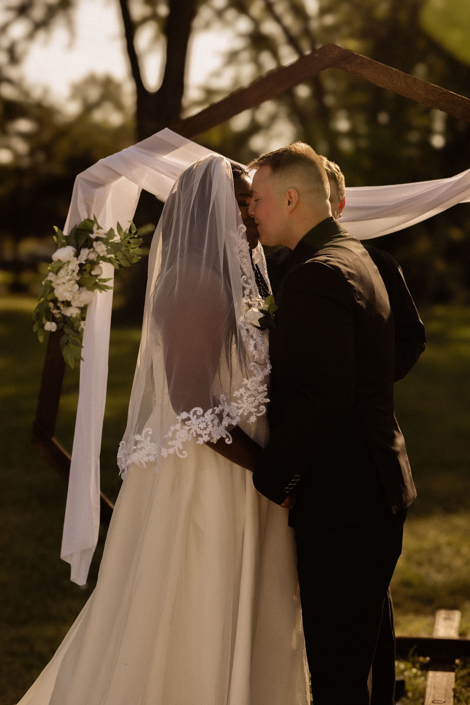 Bride and groom kiss during sunset ceremony under a white draped arch with floral decorations.