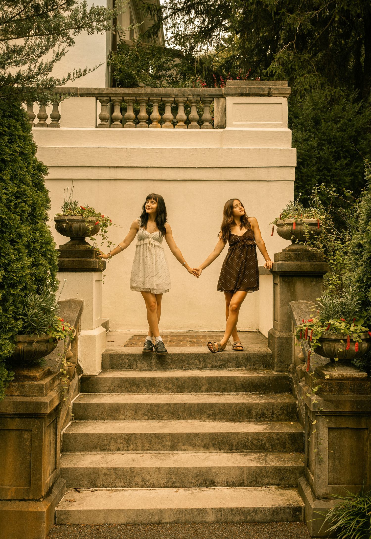 Two young women in summer dresses hold hands on elegant stone steps surrounded by lush greenery and decorative urns.