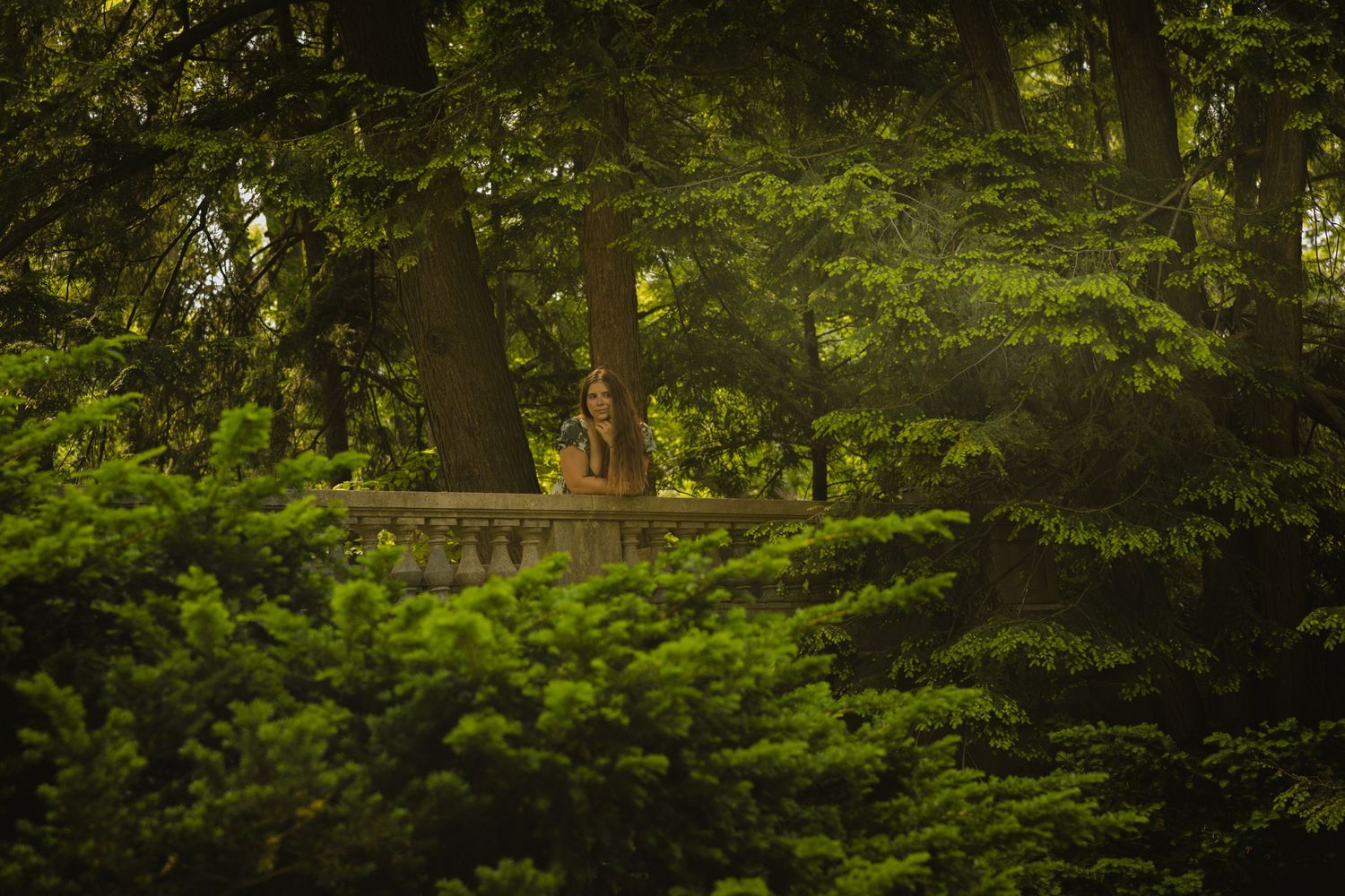 A person stands on a stone balustrade surrounded by lush green trees in a serene garden setting.