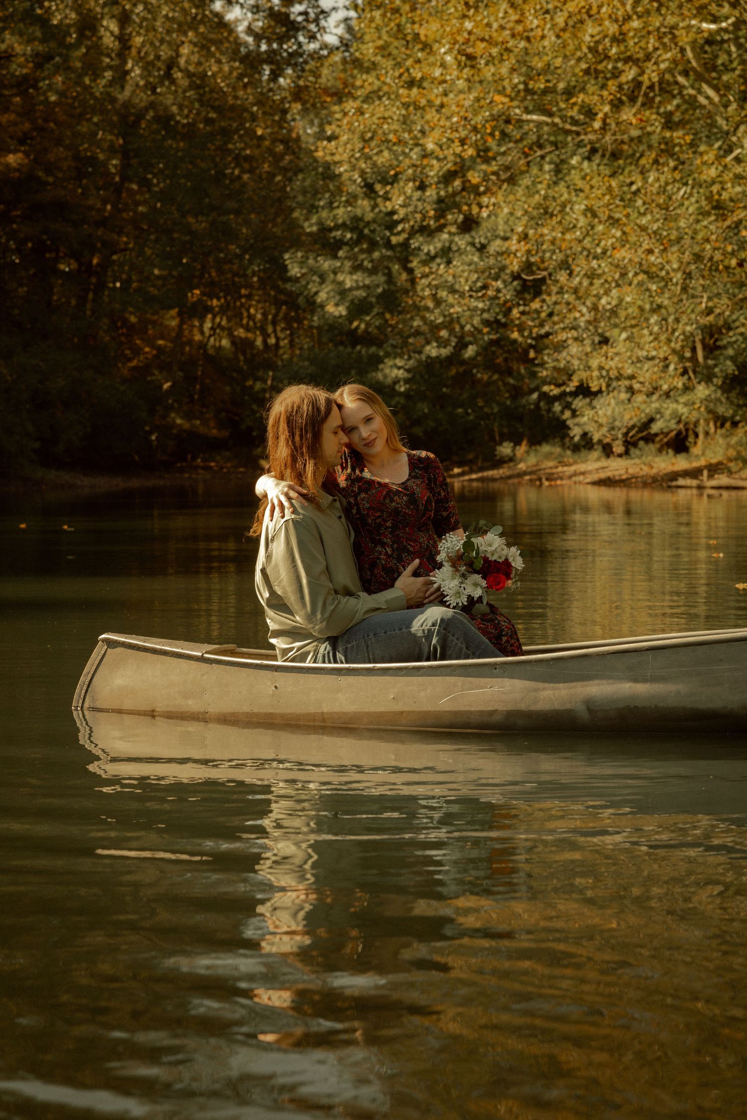 Two women share a romantic moment in a canoe on a calm river surrounded by autumn foliage, one holding a bouquet.