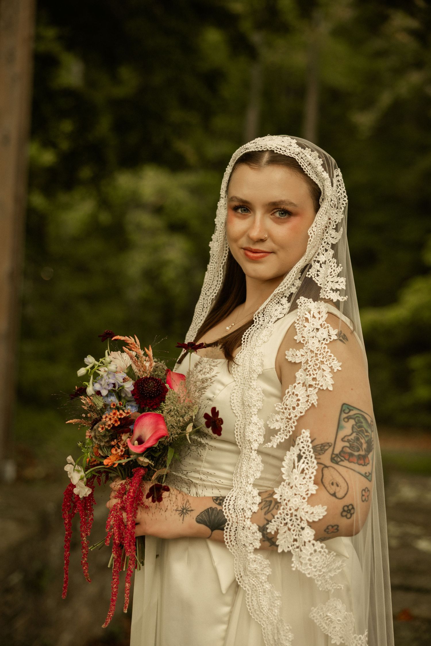 Tattooed bride in white satin gown and lace mantilla veil holds colorful bouquet outdoors.