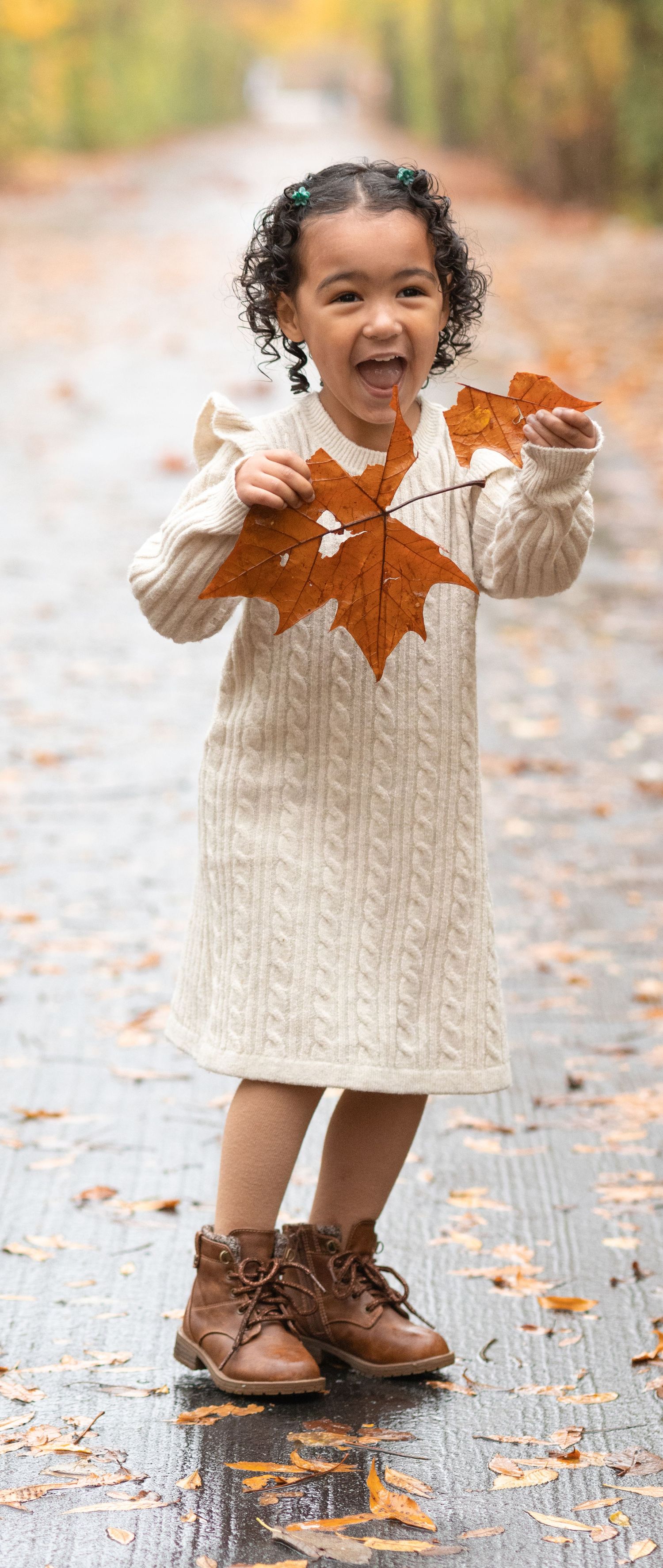 A child in a cream sweater holding autumn leaves while wearing brown boots on a wet sidewalk.