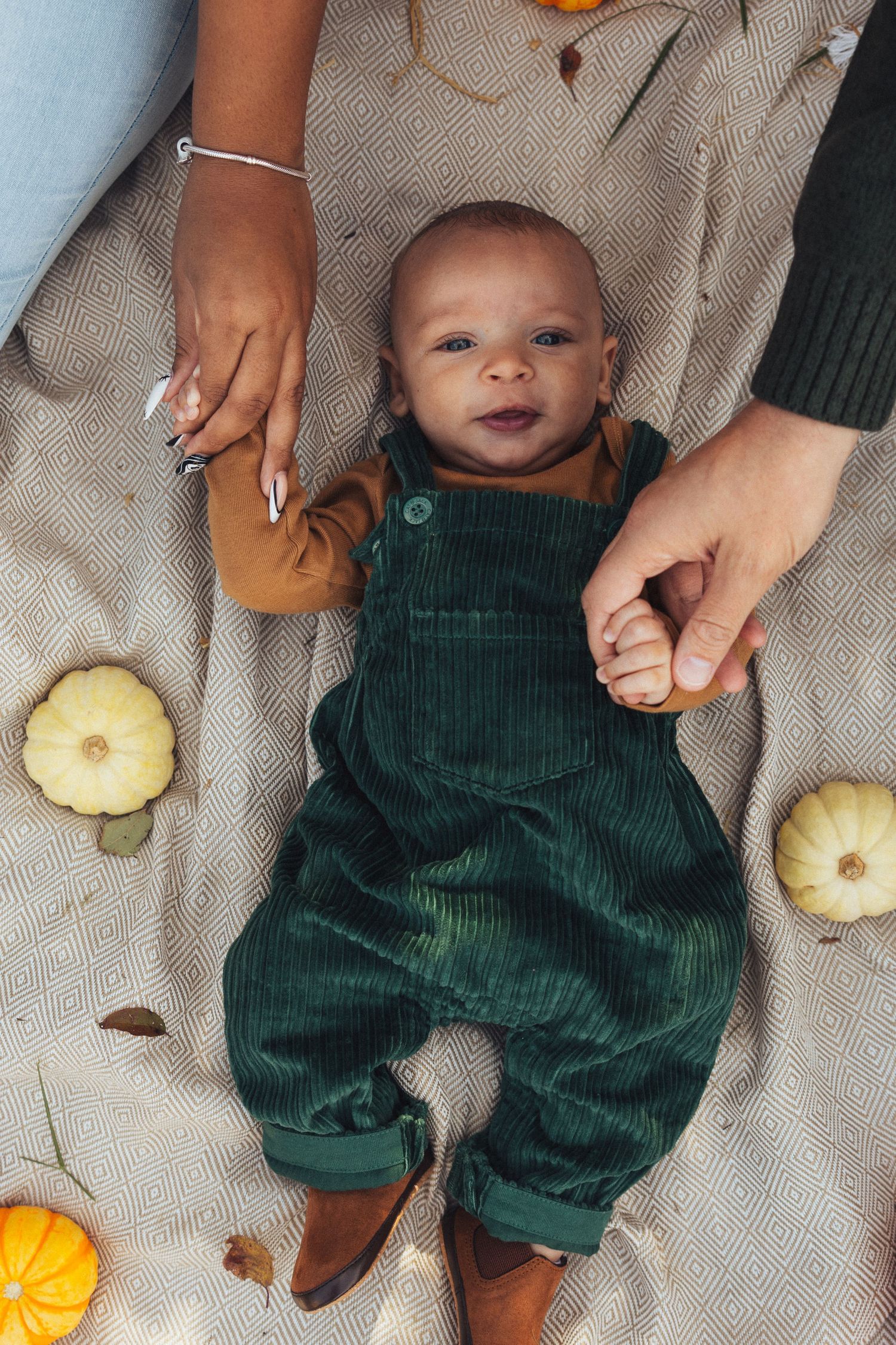 Small baby in green corduroy overalls lying on blanket with autumn gourds and decorative items.
