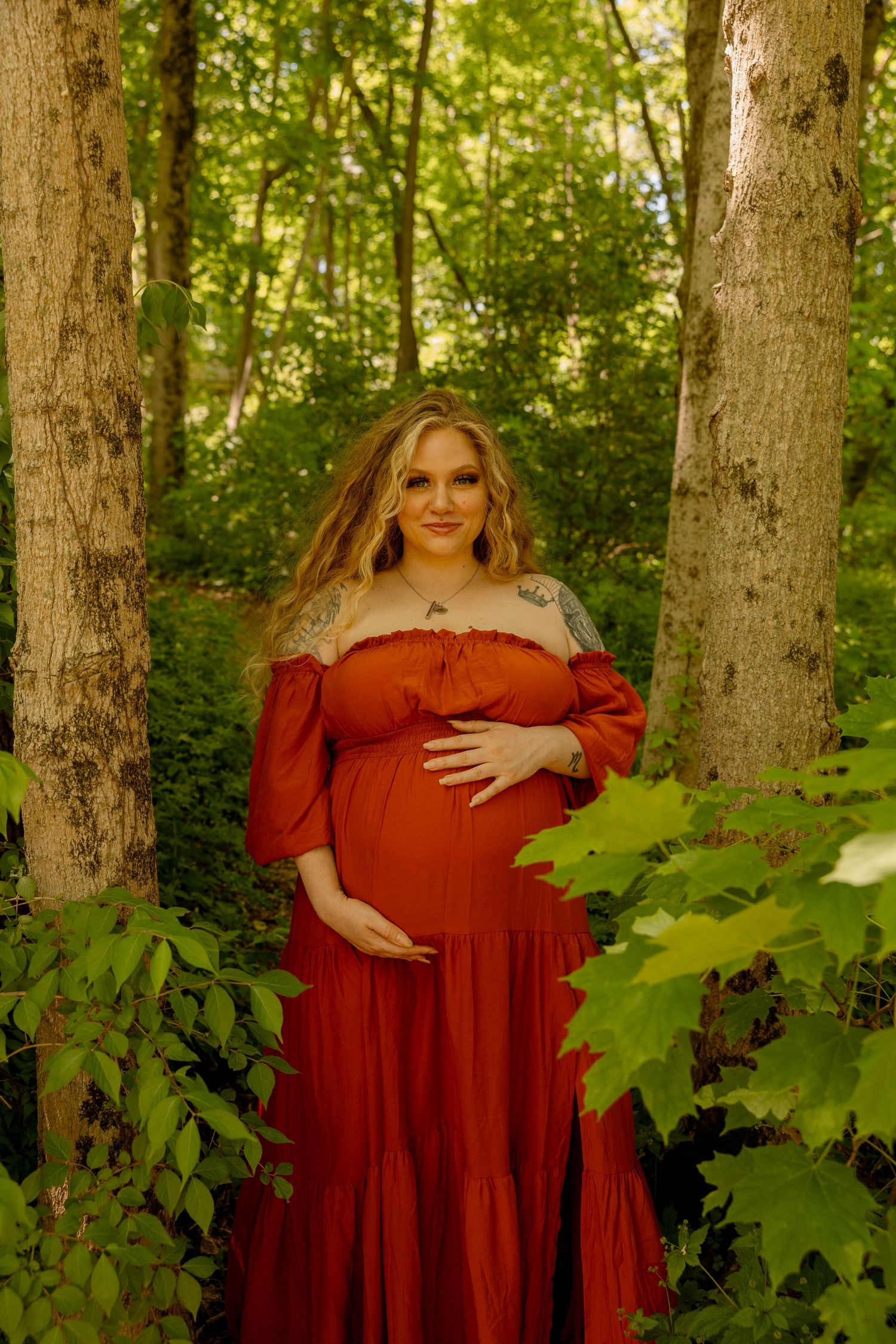 Maternity portrait in a red dress among green forest trees and maple leaves.