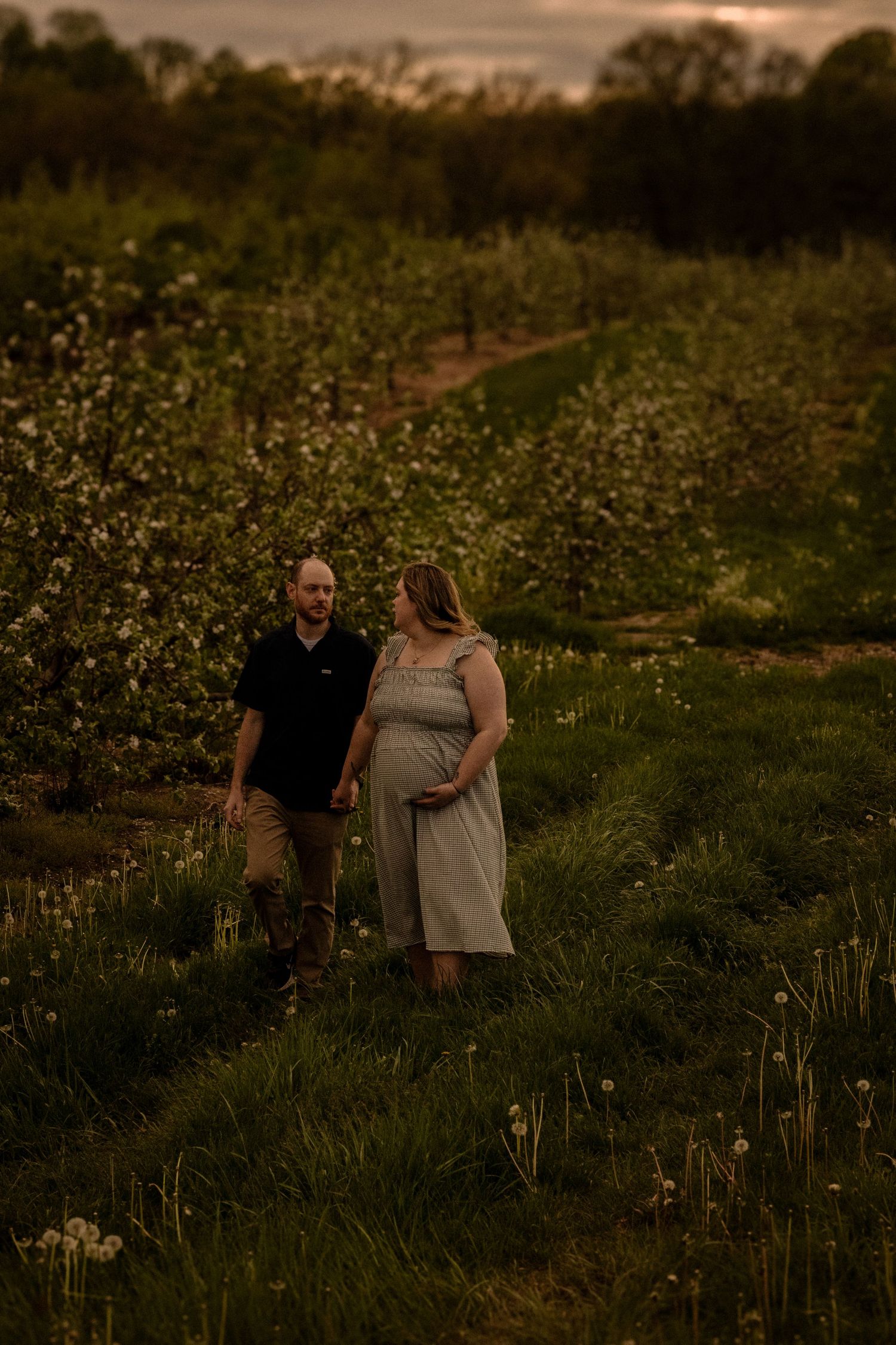Couple strolls through flowering meadow during golden hour maternity photoshoot.